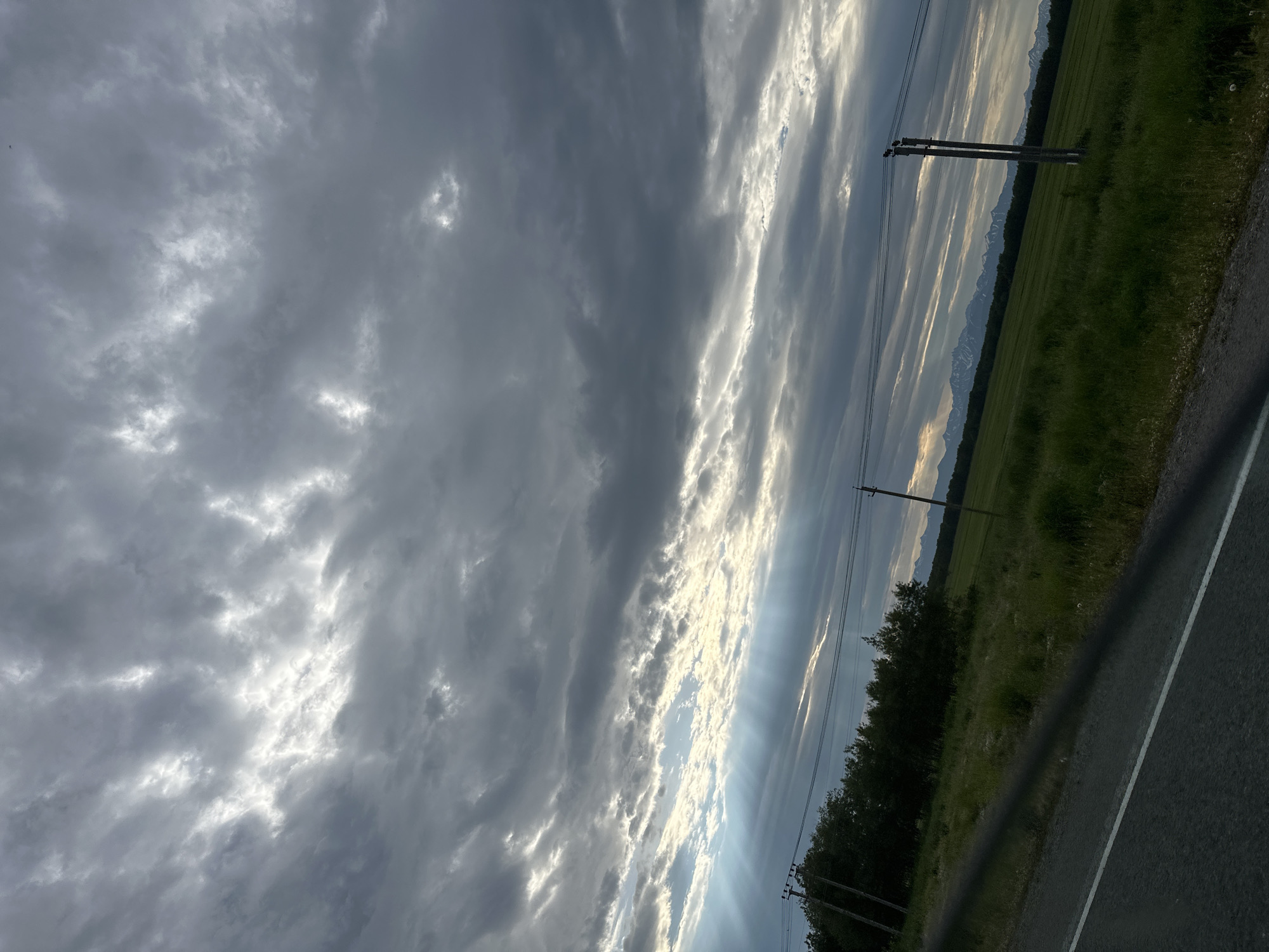 The image captures a landscape scene under a cloudy sky with visible sunbeams. In the foreground, a section of a road with a white line is visible, angled from the bottom left corner. To the right of the road, a green grassy field stretches into the distance. A line of trees borders the field on the left. In the background, mountains with snow-capped peaks are discernible. Two utility poles with wires are present in the middle ground. The sky is filled with dramatic, layered clouds, with patches of sunlight breaking through, creating shafts of light that illuminate the scene. The location is identified as Кеткино, Russia.
