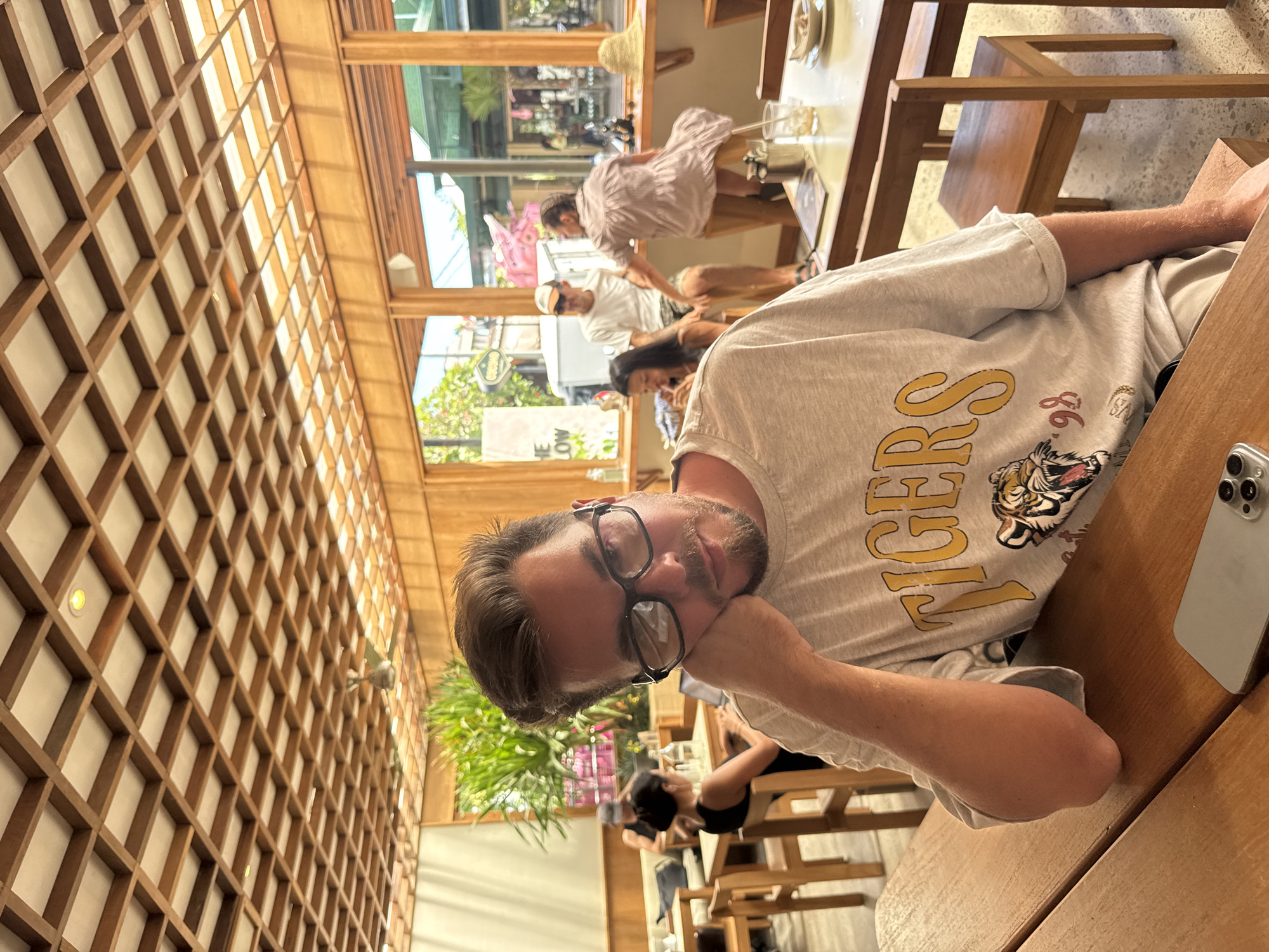 A sideways-oriented medium shot captures a man seated at a wooden table inside a restaurant with a distinctive architectural feature – a lattice-patterned wooden ceiling. The man, wearing glasses and a t-shirt emblazoned with "Tigers" and a tiger graphic, rests his face on his hand. To the right of the frame, a smartphone lies on the table.
The restaurant is populated with other patrons. In the background, multiple people are visible, including a group standing near the window and others seated at tables. The setting appears to be a city street, with building exteriors visible through the restaurant's windows. Plants also add to the restaurant's ambiance. The city and country of Unknown are unspecified.