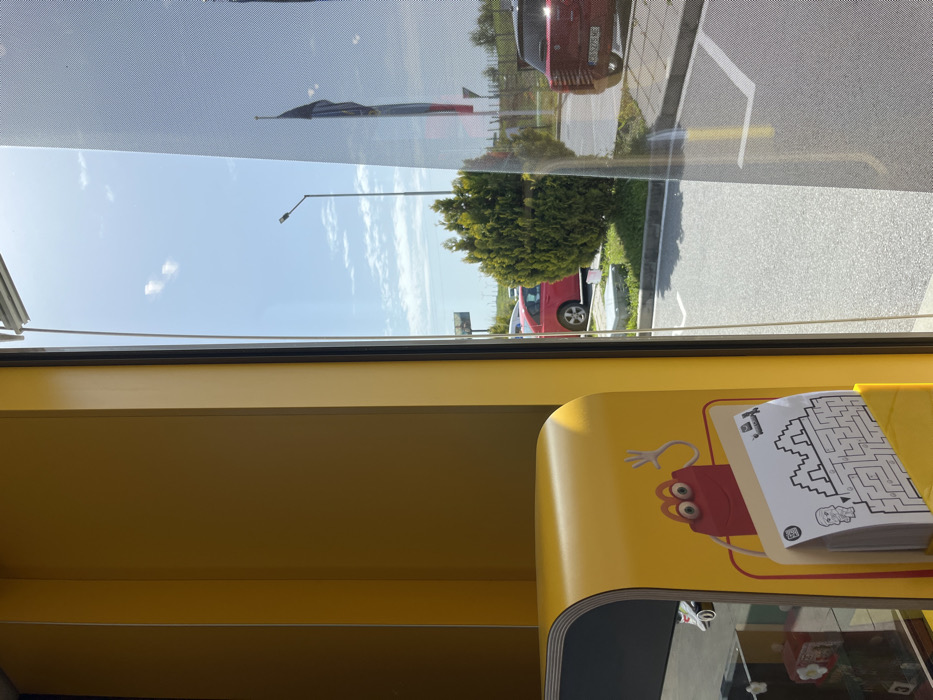 A window inside a fast-food restaurant, likely McDonald's, in Hadjidimitrovo, Bulgaria, shows a view of the parking lot. The interior is mostly yellow with a black and yellow countertop, and a cartoon figure of Ronald McDonald is visible on the front of the counter. A black and white maze printout is sitting on the countertop. The exterior view from the window shows a blue sky with a few white clouds and a streetlight. A car, likely red in color, is parked in the lot. There is a small bush in the foreground and a flagpole with a Bulgarian flag can be seen in the background. The ground is paved with gray concrete. The scene is brightly lit and there are no people visible.