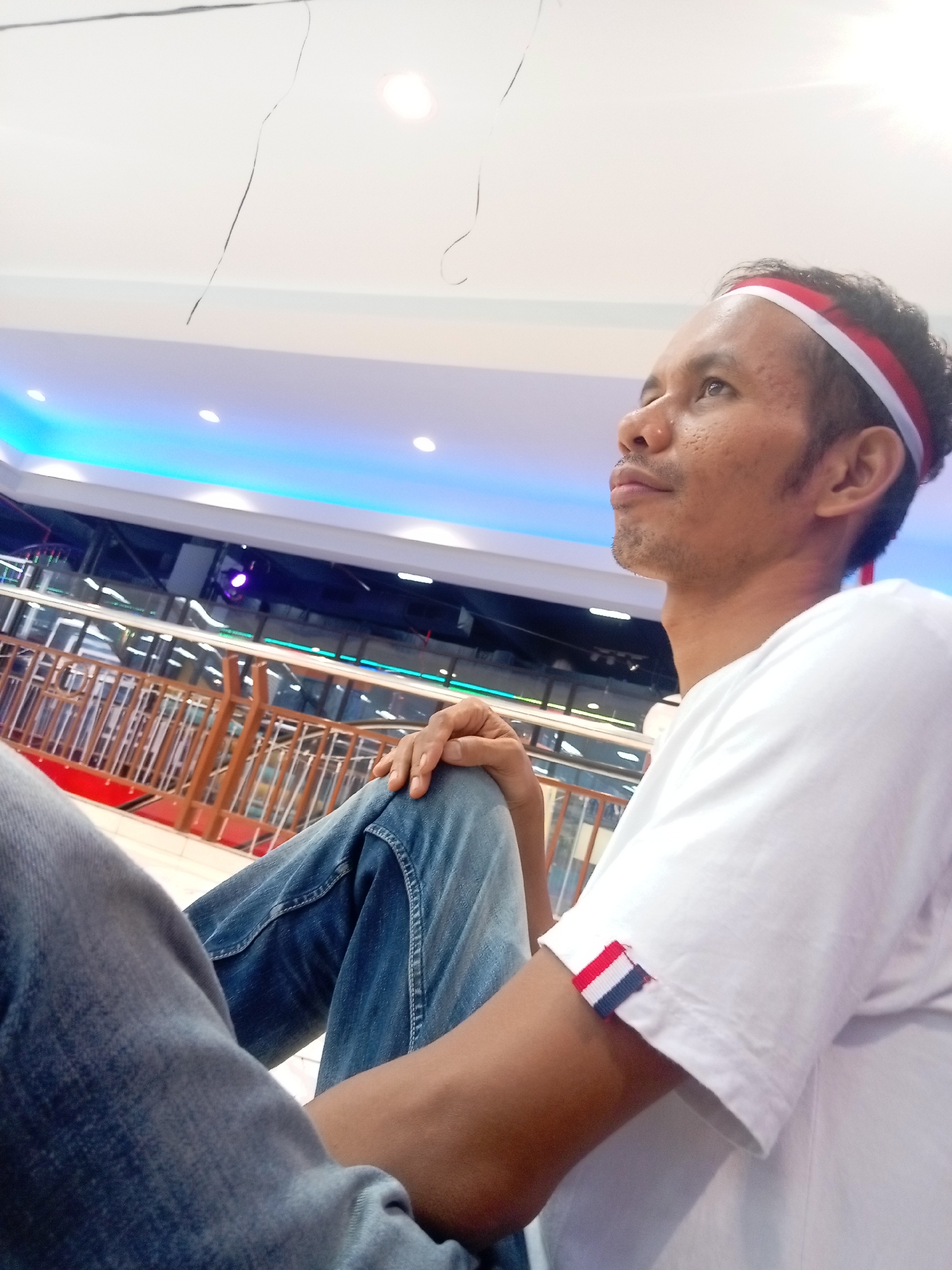 A man is sitting indoors, looking upwards and to the right. He has a red and white striped band on his forehead and a similar red, white, and blue striped band on his right sleeve. He is wearing a white t-shirt and blue jeans. His left leg is crossed over his right. In the background, there are railings and illuminated blue lights on the ceiling. The environment appears to be a public indoor space, possibly a mall or an entertainment venue. The man's posture and gaze suggest he might be watching something or someone above or in front of him. The clothing and accessories suggest a possible patriotic theme. The scene is likely in Batam, Indonesia, given the presence of red and white colors often associated with the Indonesian flag.