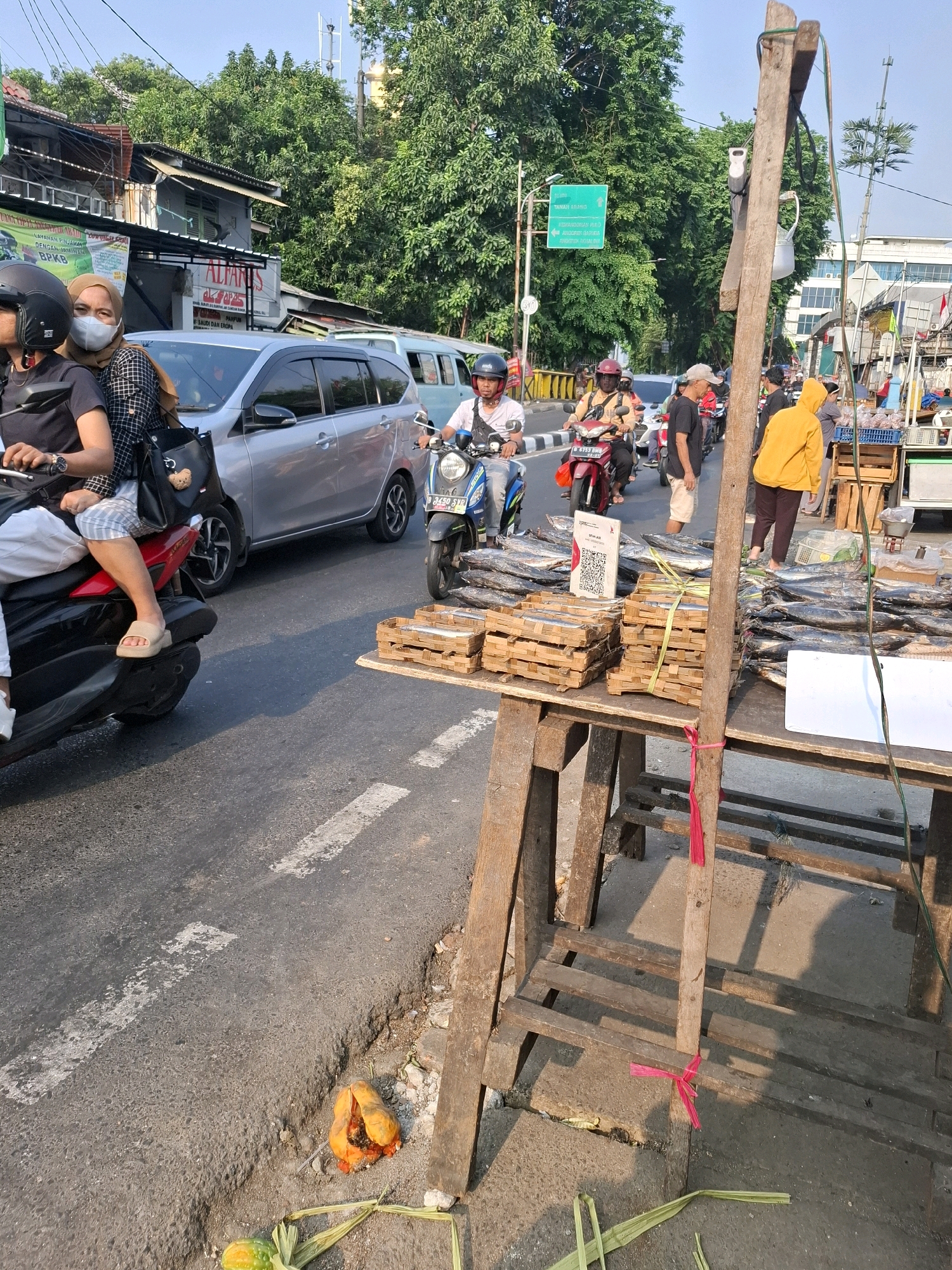 A street scene in South Jakarta, Indonesia, depicts a busy road with traffic, including motorcycles and cars. On the right side of the road, a stall is set up selling dried fish. Several stacks of dried fish are arranged on a wooden table, some of which are bundled with straw. A QR code is visible on one of the fish stacks. A wooden pole with pink ribbons tied to it stands near the stall. In the background, buildings, trees, and power lines are visible. Several people are present, some on motorcycles and others near the stall. One person in a yellow hoodie is standing by the stall, and another is walking on the sidewalk. The road is paved and marked with white lines. In the foreground, on the edge of the road, are some discarded fruits, possibly papayas, and palm leaves.