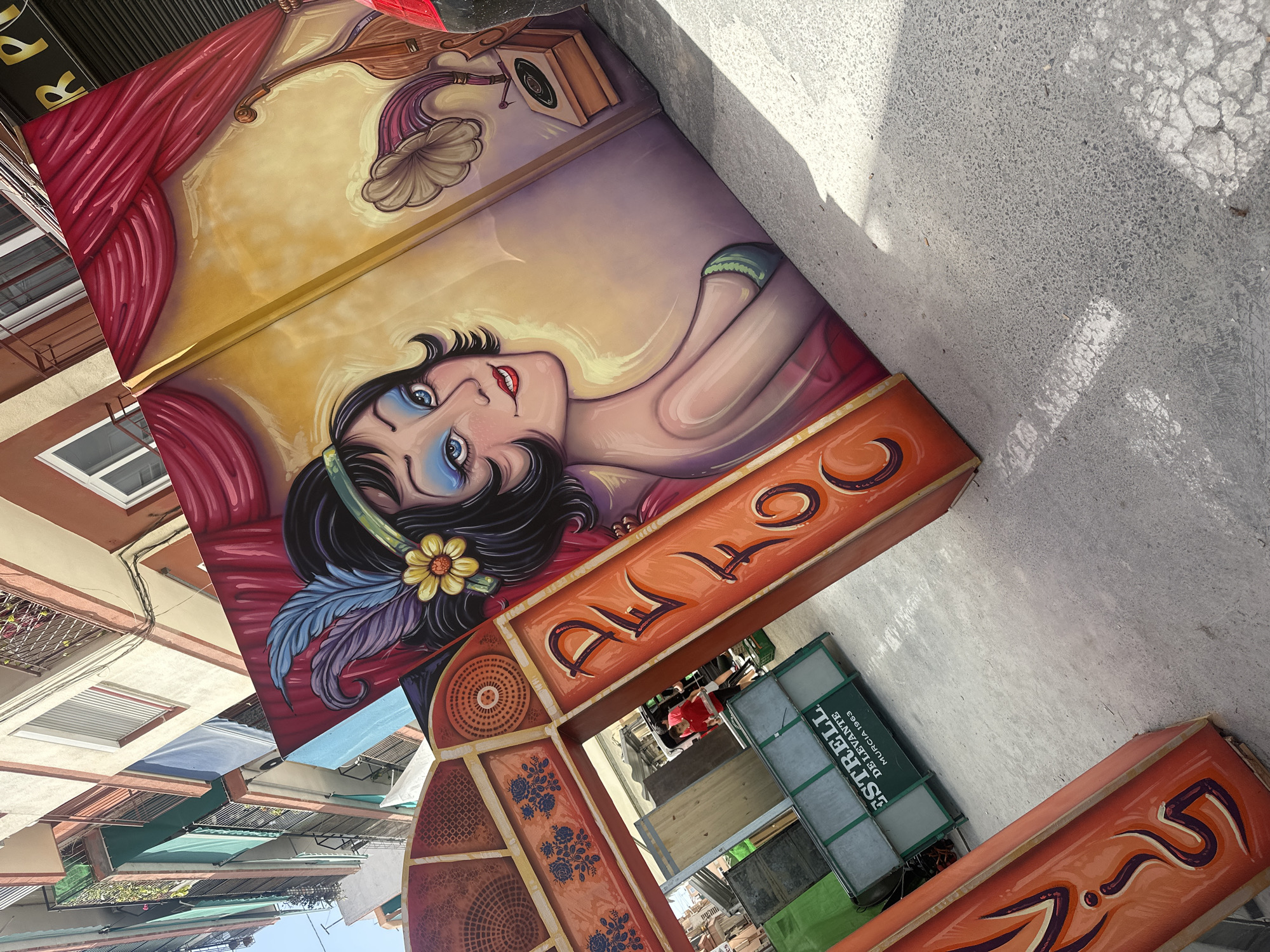 This is an outdoor, high-angle shot of a colorful, large-scale artwork displayed vertically on a street. The artwork depicts a stylized art deco portrait of a woman with dark hair, large blue eyes, and red lips. She is adorned with a yellow flower and blue and purple feathers in her hair, and wearing a red dress. Behind her, a gramophone is visible. The artwork is framed with ornate borders featuring floral patterns and swirling orange and purple designs.
Below the main portrait, a section of the artwork reveals a title in stylized orange lettering with dark purple outlines. To the right of this, a green sign with white lettering reads "ESTRELL DE LEVANTE MURCIA 1903". This sign is attached to a structure that appears to be a booth or stall, with wooden shelves and merchandise visible behind it.
The scene is outdoors, with a concrete ground surface in the foreground where shadows from sunlight are cast, indicating it's daytime with bright sun. The background shows buildings with balconies and awnings, suggesting an urban or town setting. The overall impression is one of a vibrant street art display, possibly part of a festival or market given the presence of the stall. The artwork evokes a sense of retro glamour and festivity.