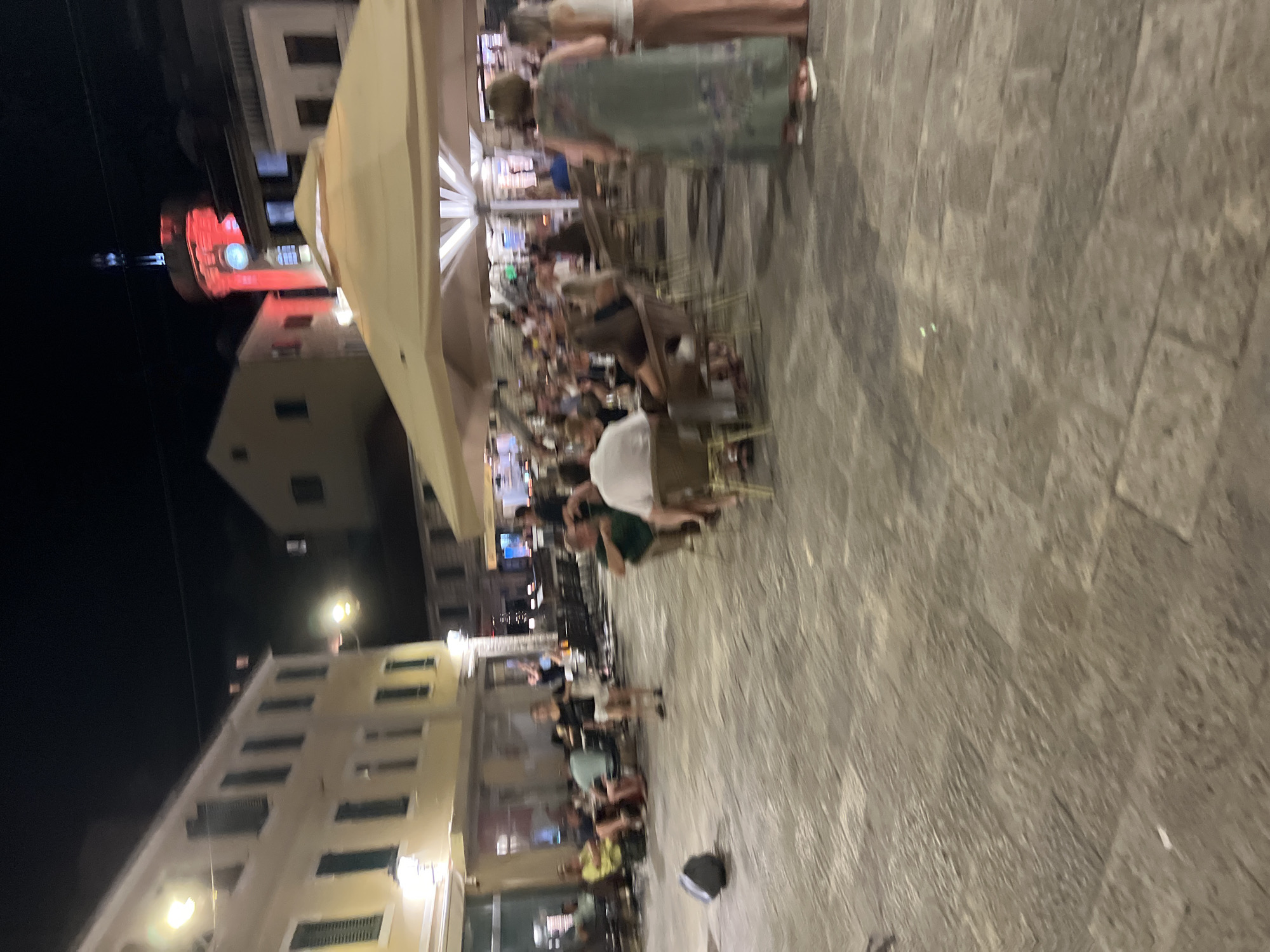 The image captures a lively outdoor scene at night in Herceg Novi, Montenegro, likely near the central square or a promenade. A large, light-colored umbrella shades a seating area where numerous patrons are gathered, dining and socializing. People are seated at tables, some appearing to be couples, others in small groups. The architecture of the surrounding buildings, illuminated by streetlights and some with red accents on a tower, suggests a historic European town. The ground is paved with large stone slabs. In the foreground, a dark, cylindrical object, possibly a speaker or a bin, rests on the pavement. The atmosphere appears relaxed and social, with evidence of a bustling evening in a public space.