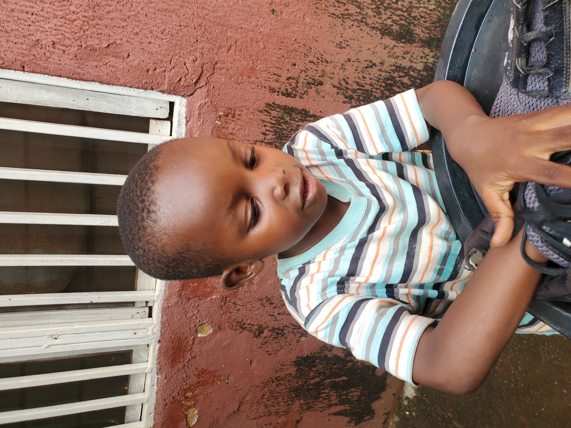 A young boy is shown in a close-up shot, positioned on his side. He is wearing a striped t-shirt with alternating bands of white, light blue, dark blue, and orange. His eyes are closed, and his facial expression suggests a state of relaxation or perhaps sleep.
The background consists of a reddish-brown textured wall, partially covered with some green mossy patches, indicating an outdoor setting. To the left, a white window grill with horizontal bars is visible. A dark, round object, possibly a basin or container, is present on the right side of the frame, and the boy's right arm is resting inside or near it, with a black object, likely a shoe, also visible.
The lighting suggests it could be daytime, but the specific time is not discernible. The presence of moss on the wall and the textured concrete might indicate a somewhat humid or weathered environment. No text is visible in the image. The overall impression is a candid moment captured of a child in a simple outdoor environment in Jos, Nigeria.