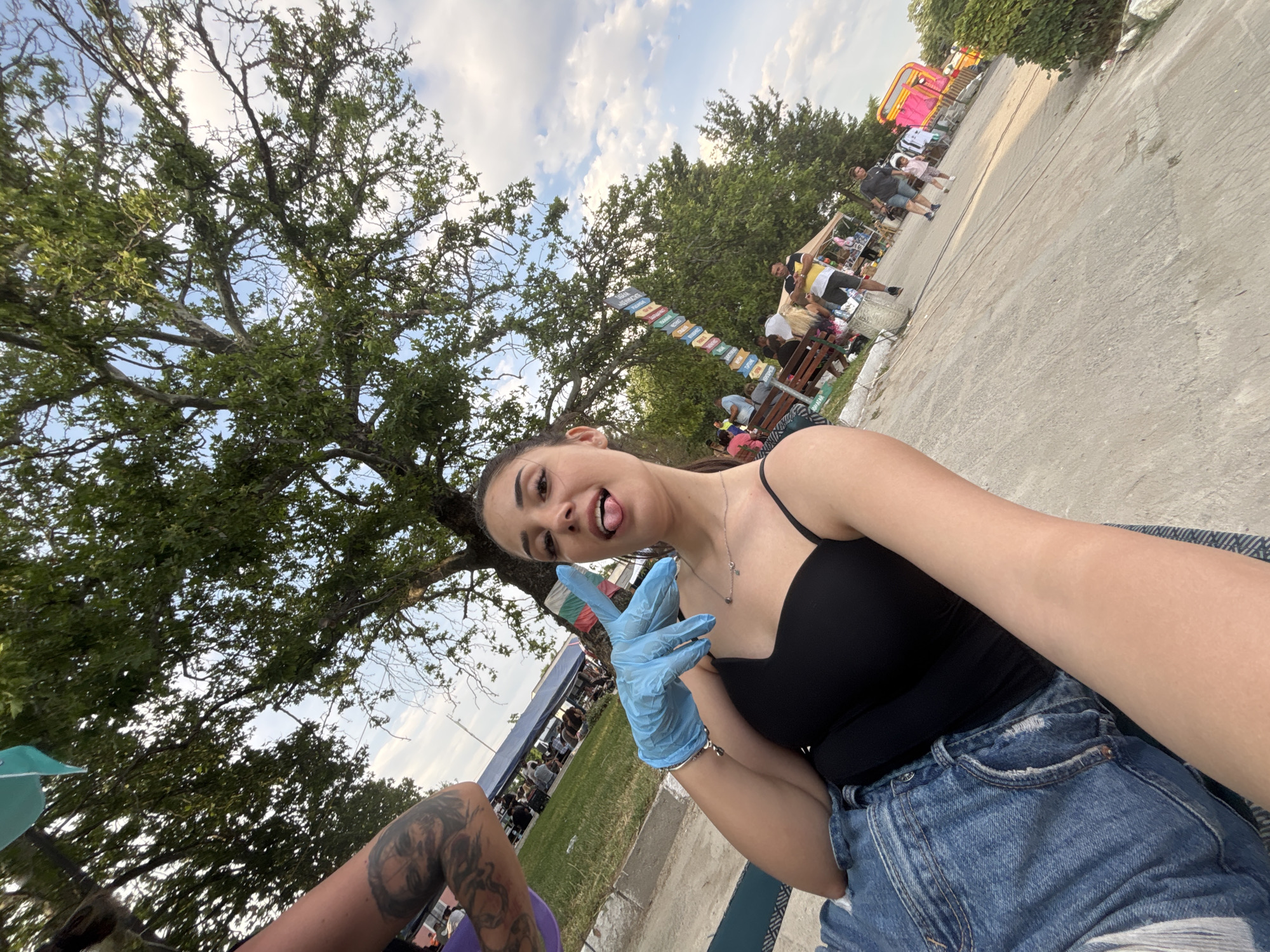 The image depicts a low-angle, wide-angle outdoor selfie featuring a young adult female in the foreground, situated in Kostievo, Bulgaria.
The primary subject, a light-skinned female with dark hair pulled back, occupies the lower-left to mid-right of the frame. She wears a black spaghetti strap top and blue denim shorts. A silver-colored necklace with small charms is visible on her neck. Her left hand, covered by a light blue disposable glove, is raised, forming a "V" or peace sign with her index and middle fingers. Her tongue is extended from her mouth. Part of her right upper arm, adorned with a detailed tattoo, is visible in the upper-left corner, suggesting another individual is seated closely beside her. She appears to be seated on a textured mat or patterned surface.
The mid-ground to background reveals an outdoor event or gathering. To the left, a blue tent structure is visible, surrounded by several individuals sitting and standing. A Bulgarian flag (white, green, red stripes) is partially visible near this tent. Large, mature trees with dense green foliage dominate the upper portion of the image, providing shade. The sky above is partly cloudy with patches of blue.
To the right, a paved pathway extends into the distance. A multi-directional signpost with numerous colored signs is positioned near the trees. Several people are scattered along the pathway, including a man in a yellow and black striped shirt near a market stall, and further down, a man walking with a small child. In the far right background, a bright pink inflatable structure, possibly a bouncy castle, is discernible. The overall scene suggests a local fair, market, or community event taking place on a bright day.