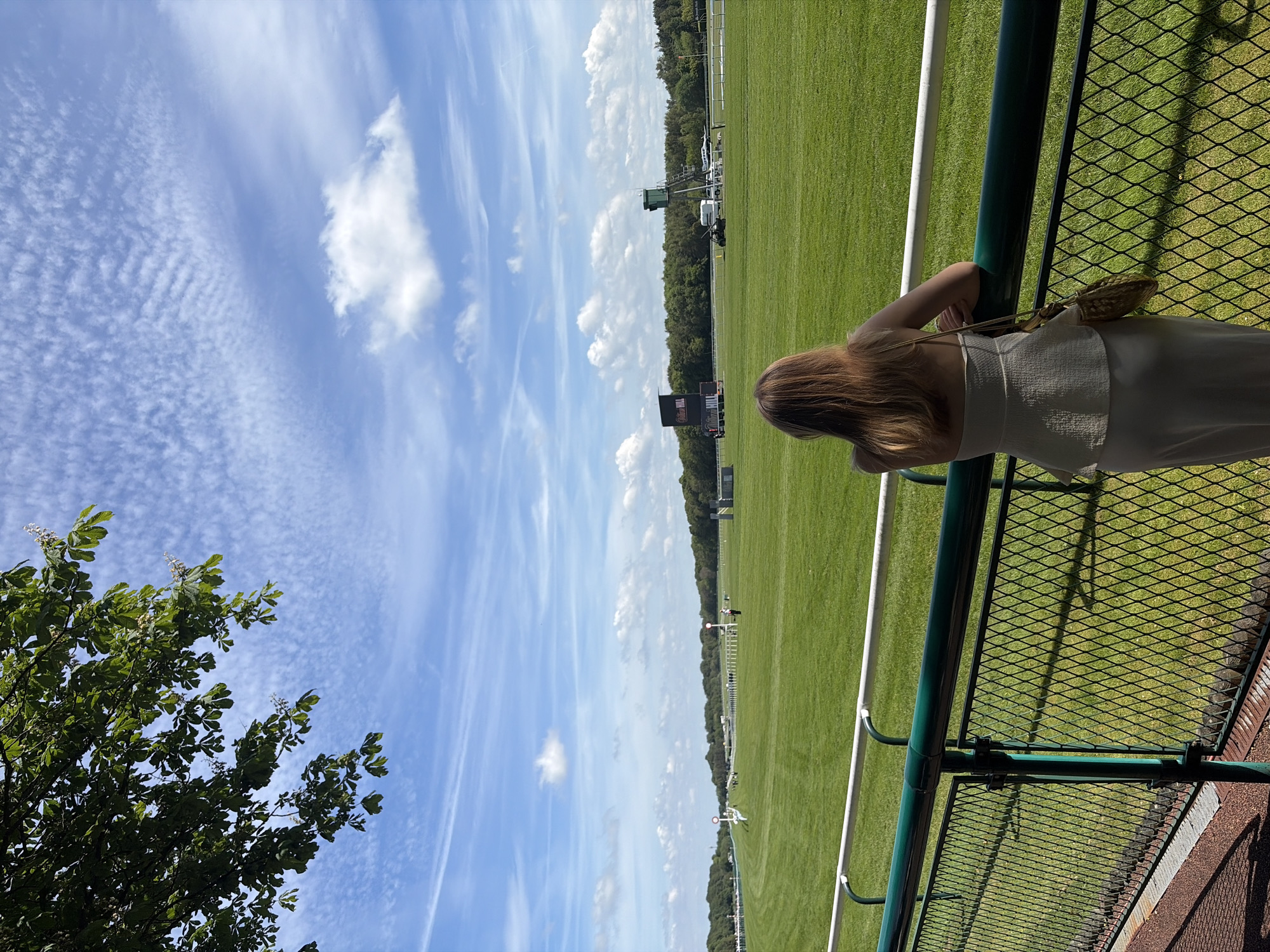 A woman with long, light brown hair is standing behind a green metal railing, looking out over a large green field on a sunny day. The field appears to be a sporting ground, possibly for polo or a similar equestrian sport, with white markers delineating the boundaries. Beyond the field, a line of trees forms a natural backdrop. In the distance, a scoreboard and some vehicles are visible. The sky is blue with scattered white clouds. The woman is wearing a white top and light-colored pants. A tan handbag hangs from her shoulder. Green trees are visible on the left. The location is Unknown, Unknown.