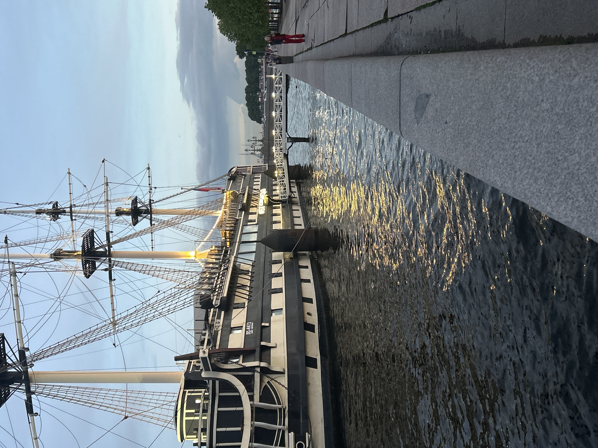 This image depicts a large, dark-hulled sailing ship docked alongside a stone embankment on a body of water. The ship features multiple masts with intricate rigging and is illuminated by warm lights. A white pedestrian bridge extends from the ship to the embankment. On the embankment, a person in red pants and a dark jacket walks away from the camera. The sky is a soft blue with scattered clouds, suggesting it is either early morning or late evening. Trees line the far side of the embankment, and distant buildings are visible. The water reflects the ship's lights, creating shimmering patterns. The scene appears to be a waterfront promenade in a city.