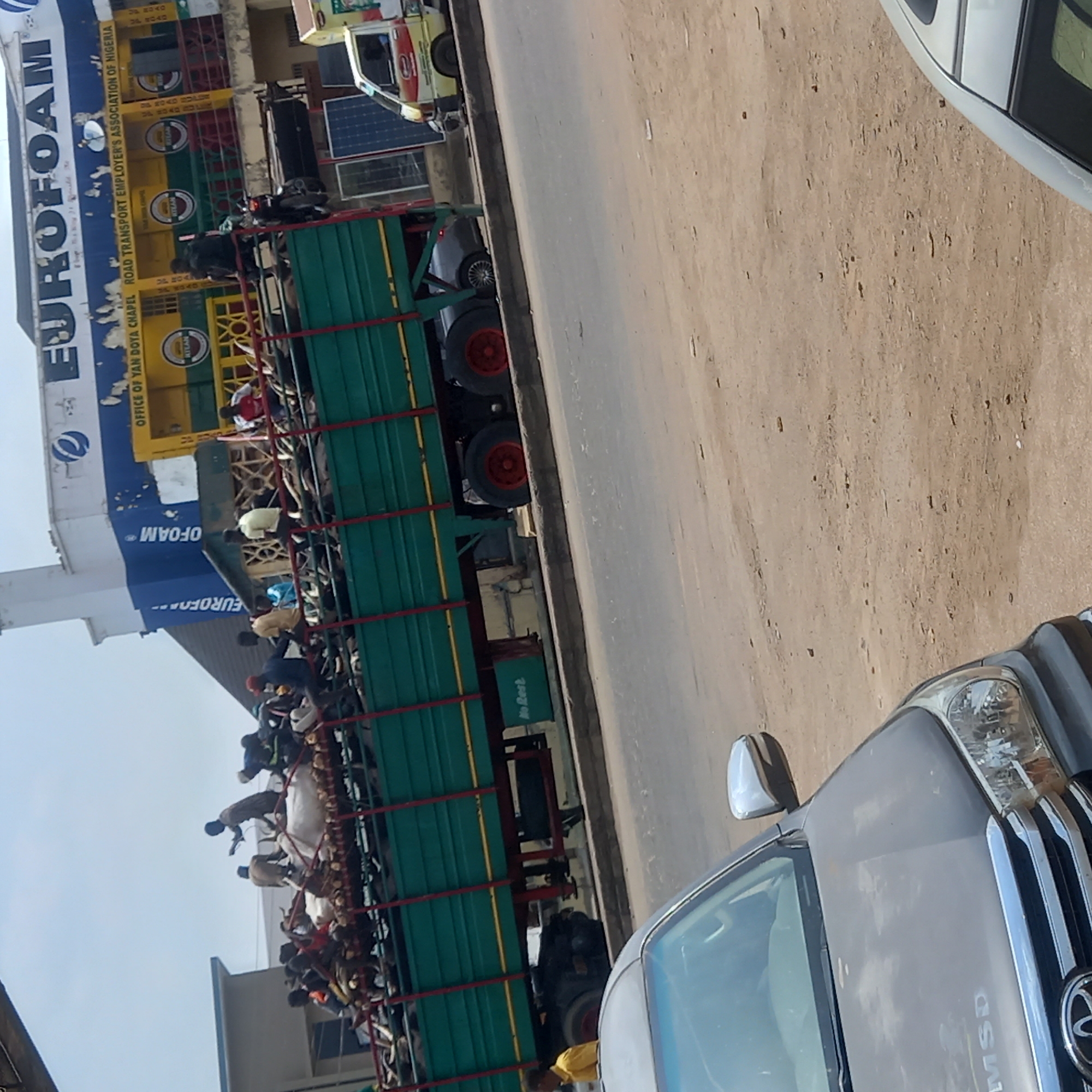 A horizontal-angle daytime shot reveals a bustling street scene in Dadi Riba, Nigeria.
On the left, a large green truck, filled with many visible people, dominates the scene. The truck also bears an indication of "No Rest".
Adjacent to the truck, is a building with "EUROFOAM" prominently displayed, as well as a yellow and green sign with "OFFICE OF YAN DOYA CHAPEL ROAD TRANSPORT EMPLOYER'S ASSOCIATION OF NIGERIA".
In the foreground, the hood of a grey vehicle is visible, positioned in the lower right. The vehicle bears the mark "MSD".
The street surface is cracked and uneven. Other vehicles, including a van, are visible in the background.