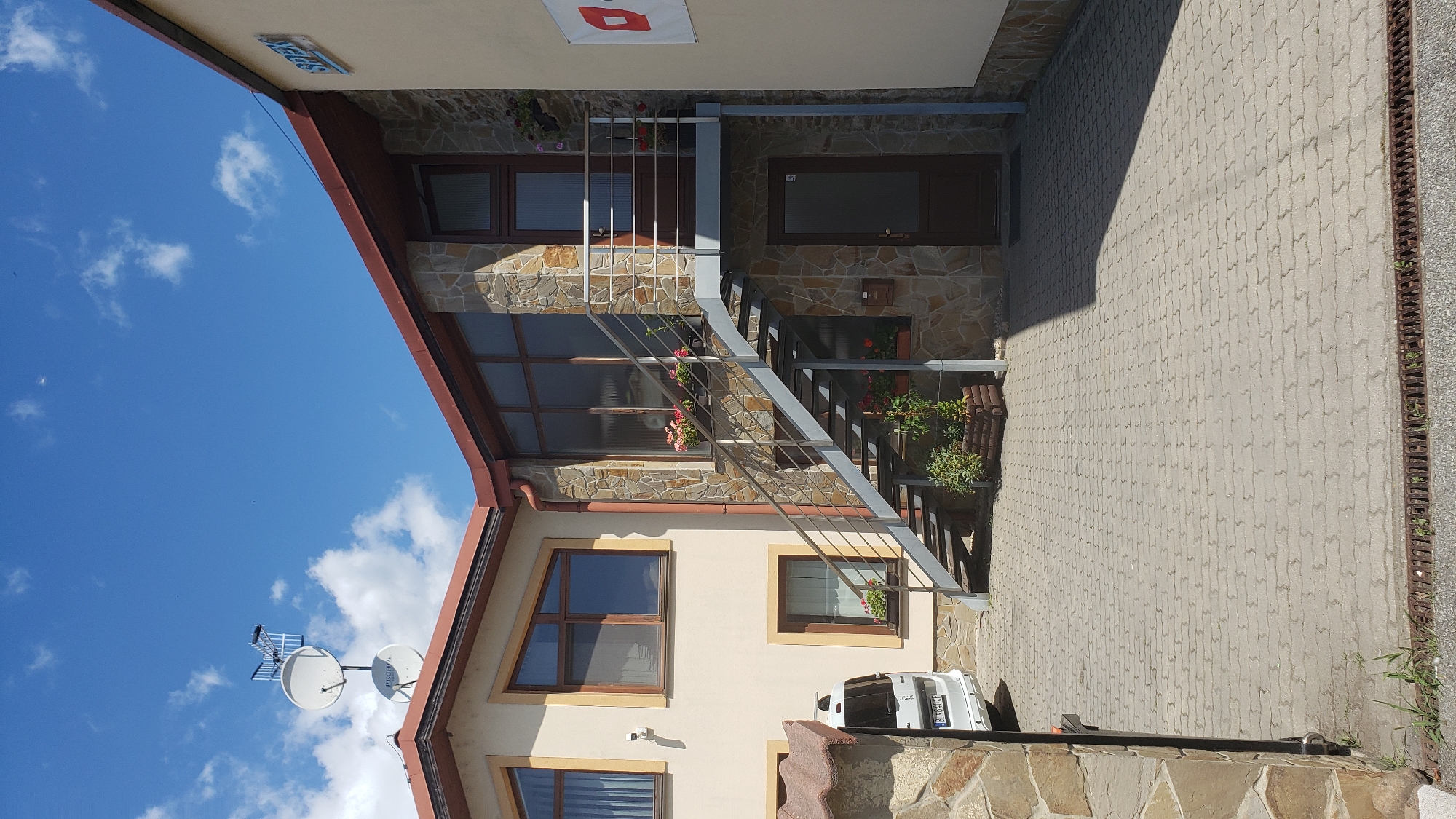 The image shows the exterior of a modern building in Prešov, Slovakia, featuring a stone facade and a metal staircase leading to an upper entrance. The building has a light-colored exterior with several windows, some with wooden frames. On the roof, there are two satellite dishes and an antenna. In front of the building, a paved courtyard is visible, with a white van parked to the left. The sky is blue with scattered white clouds. The building appears to be a commercial or residential property.