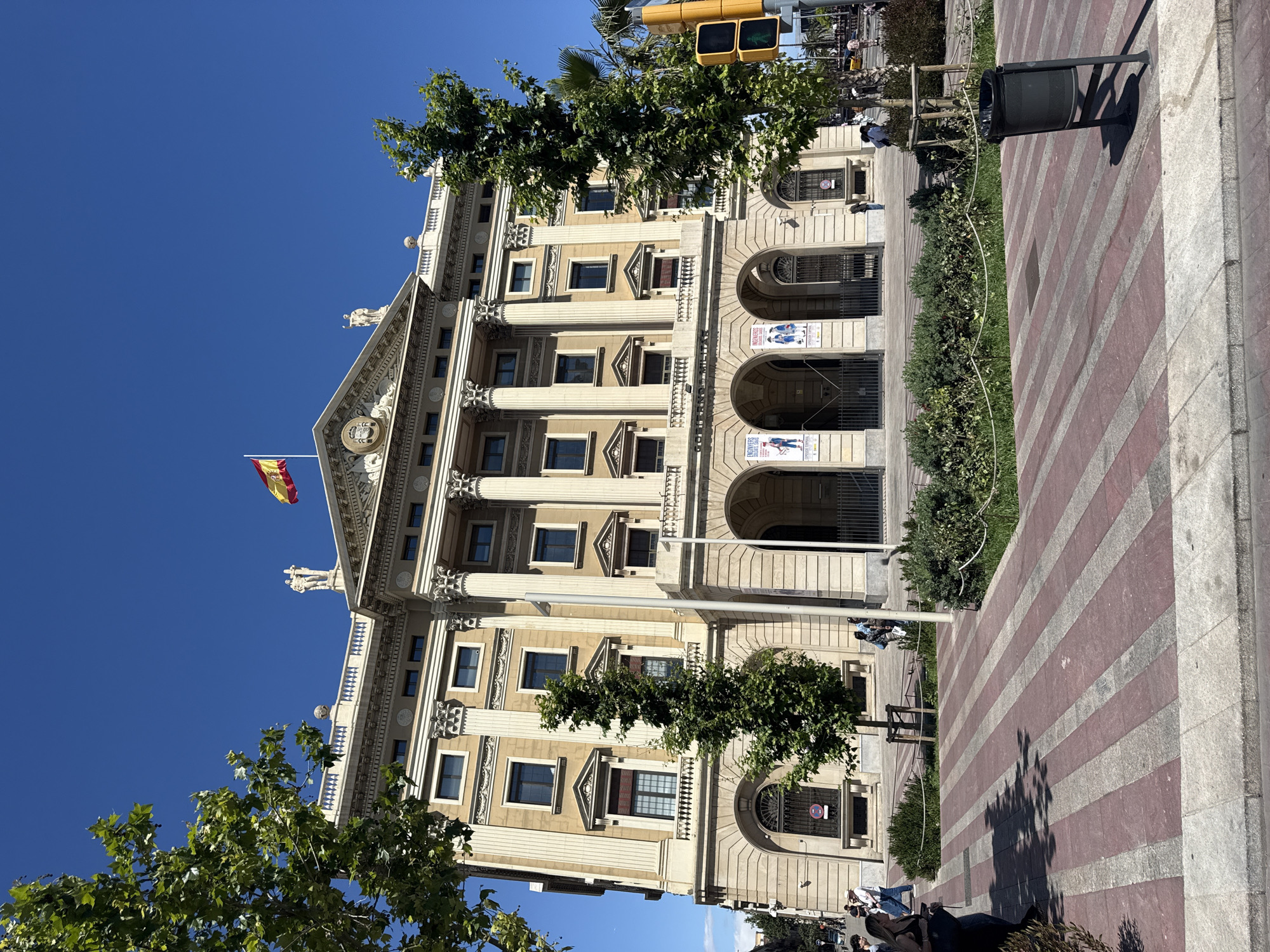 The image depicts a large, neoclassical building in Barcelona, Spain. The building has a tan facade with white columns, arched windows and doorways on the ground floor, and smaller windows on the upper floors. A Spanish flag is flying from the top of the building. The sky is clear and blue. In front of the building, there is a sidewalk with red and gray striped paving stones, separated from the building by a narrow strip of greenery. A black trash can is located on the sidewalk. Pedestrians can be seen in the foreground and along the sidewalk near the building. There are trees in the foreground, partially obscuring the view of the building. A traffic light is visible in the background.