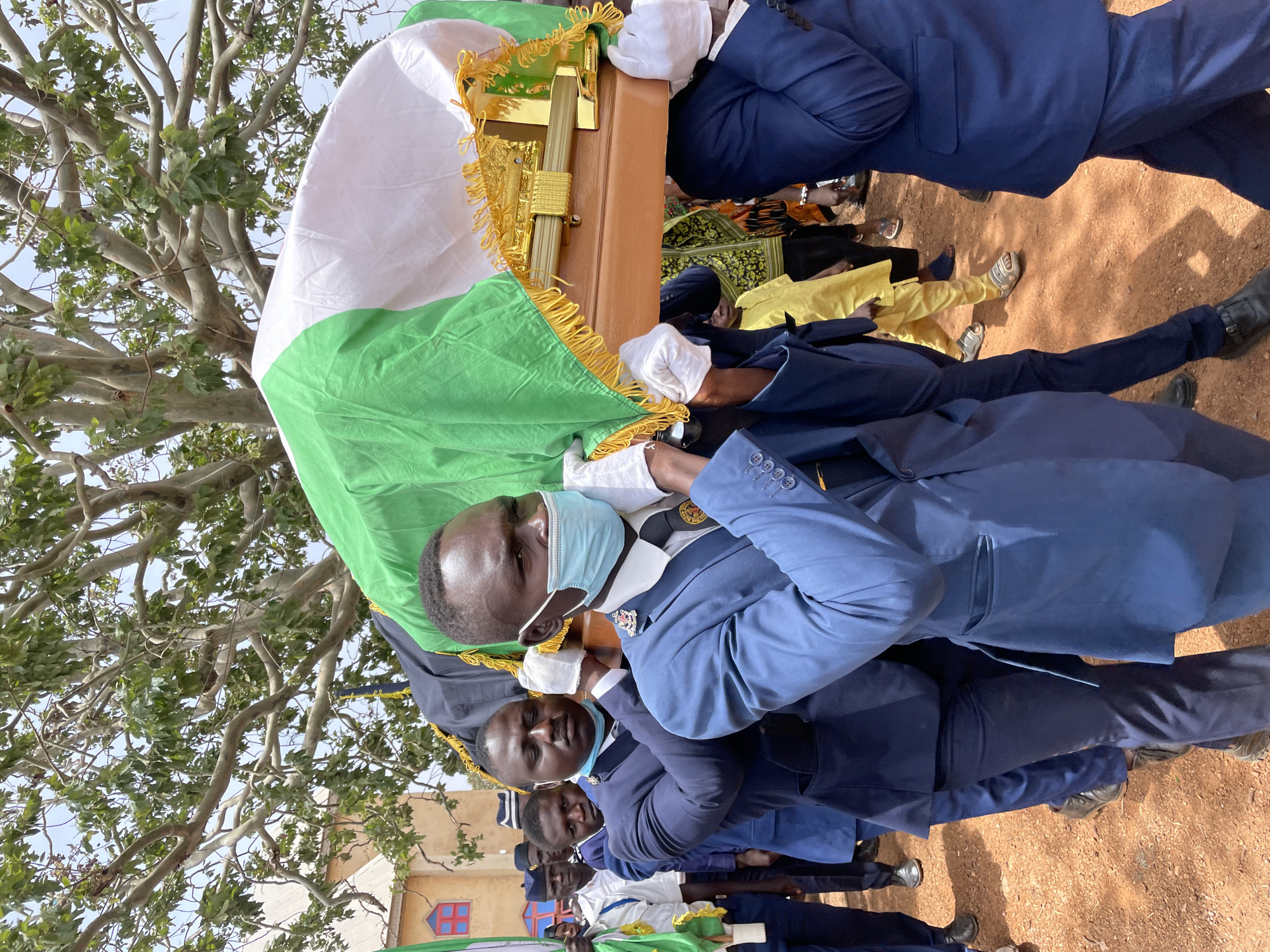 A group of men wearing light blue military uniforms are carrying a wooden coffin draped with the Nigerian flag. The men in the foreground wear light blue surgical masks. The ground is red. Some are wearing black shoes. The location is a red dirt area in Ladura Foron, Nigeria. A tree is on the left. More people stand behind the coffin carriers. Some onlookers wear yellow clothing.