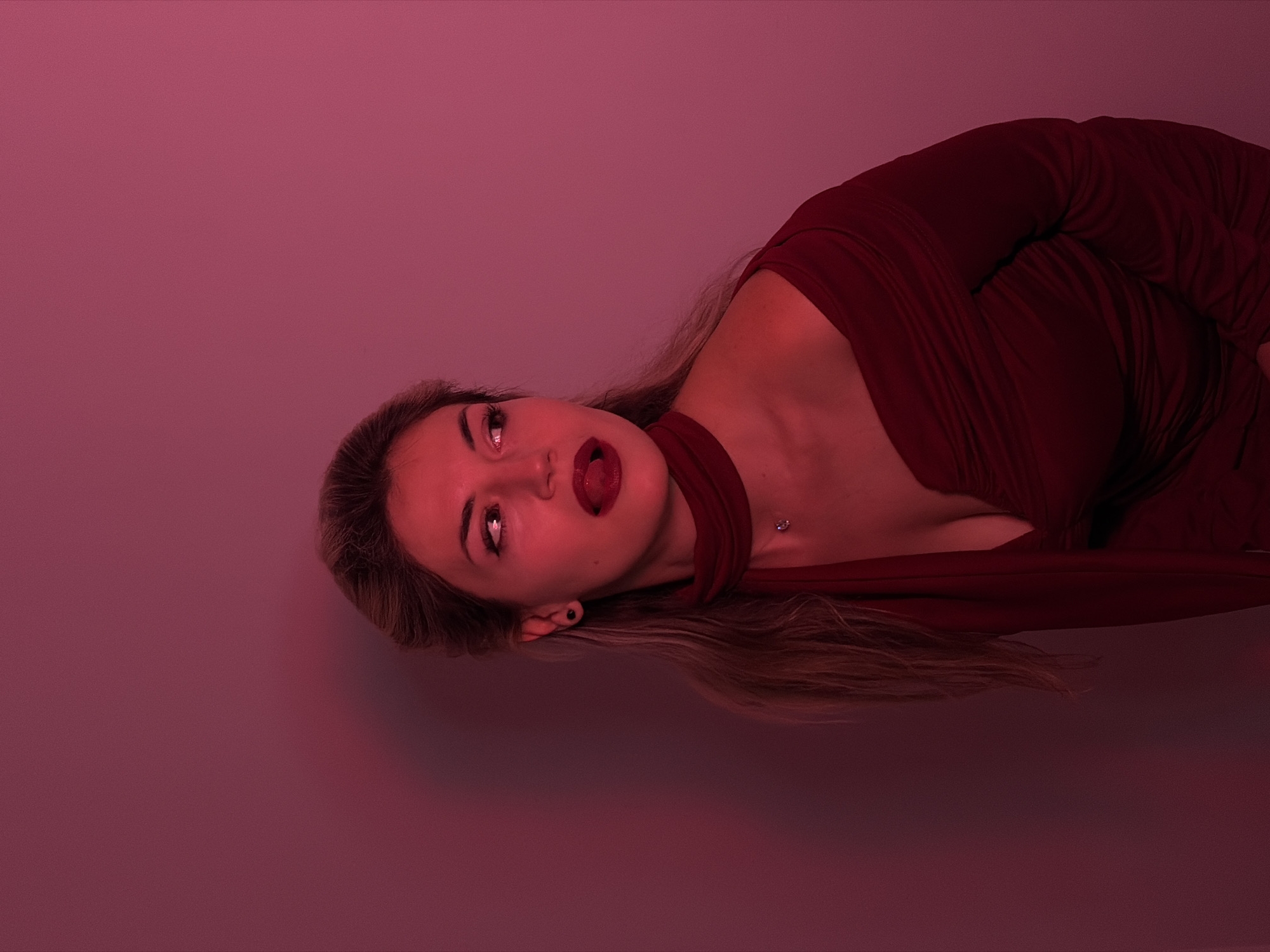A woman with long blonde hair is reclining in a magenta-lit setting. She wears a deep red dress, with part of the fabric wrapped around her neck. Her head is tilted back and to the side, and she has her tongue sticking out. The setting appears to be a solid magenta backdrop. There is no evidence to suggest this image was taken in Budva, Montenegro.