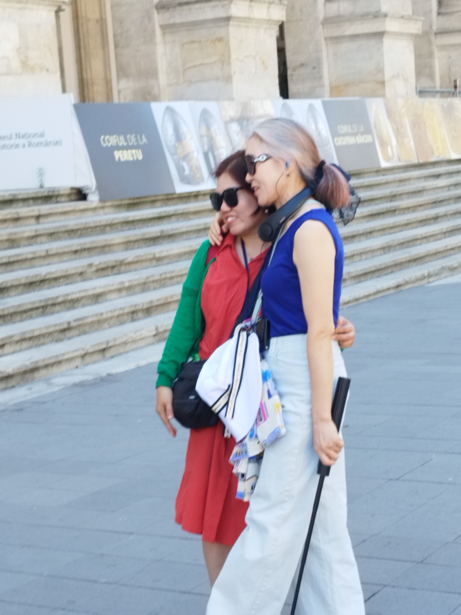 Two women are standing on a paved walkway in front of a building with stone steps. The woman on the left is wearing a red dress and a green cardigan, with sunglasses and her arm around the other woman. The woman on the right has light-colored hair, wears sunglasses, and a blue top with light-colored pants. She is holding a cane and a bag. In the background, there are signs with text in Romanian, indicating this location is likely near the National Museum of Romanian History in Bucharest, Romania.