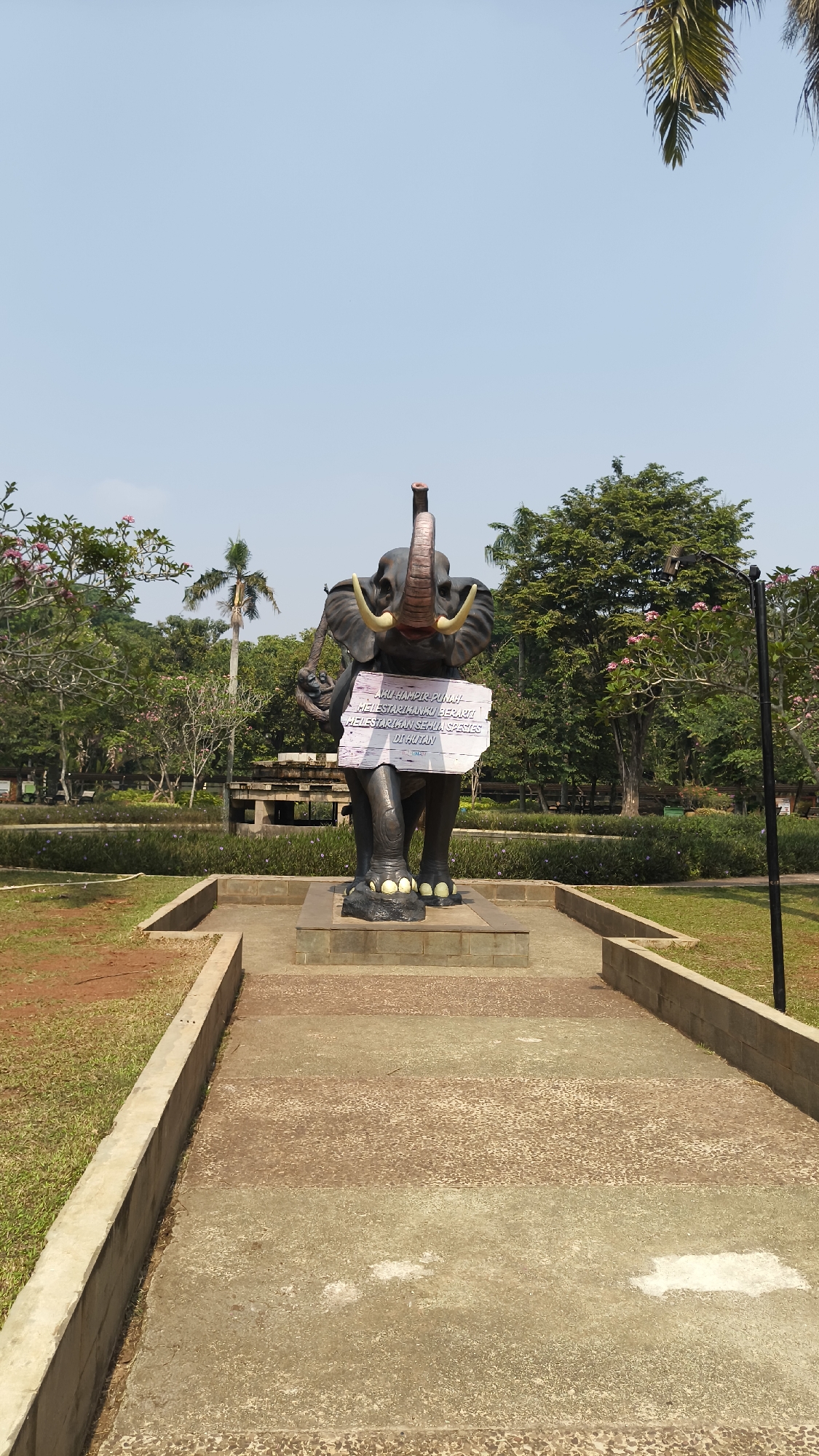The image shows a statue of an elephant in a park-like setting. The elephant statue is dark gray and stands on a pedestal. A light-colored sign with Indonesian text is attached to the elephant's chest. The sign reads: "AKU HAMIPIR PUNAH! MELASTIKANKU BERARTI MELASTIKAN SEMUA SPECIES DI HUTAN." This translates to: "I am almost extinct! Conserving me means conserving all species in the forest."
The elephant statue is surrounded by a paved pathway and a grassy area with some scattered reddish-brown dirt. In the background, there are numerous trees, some with pink flowers, and a clear blue sky. A lamppost is visible to the right of the elephant. The overall environment suggests a public park or zoo.
This statue is located in Taman Suropati, a public park in Menteng, South Jakarta, Indonesia. The park is known for its statues and is a popular recreational spot for residents of the city.