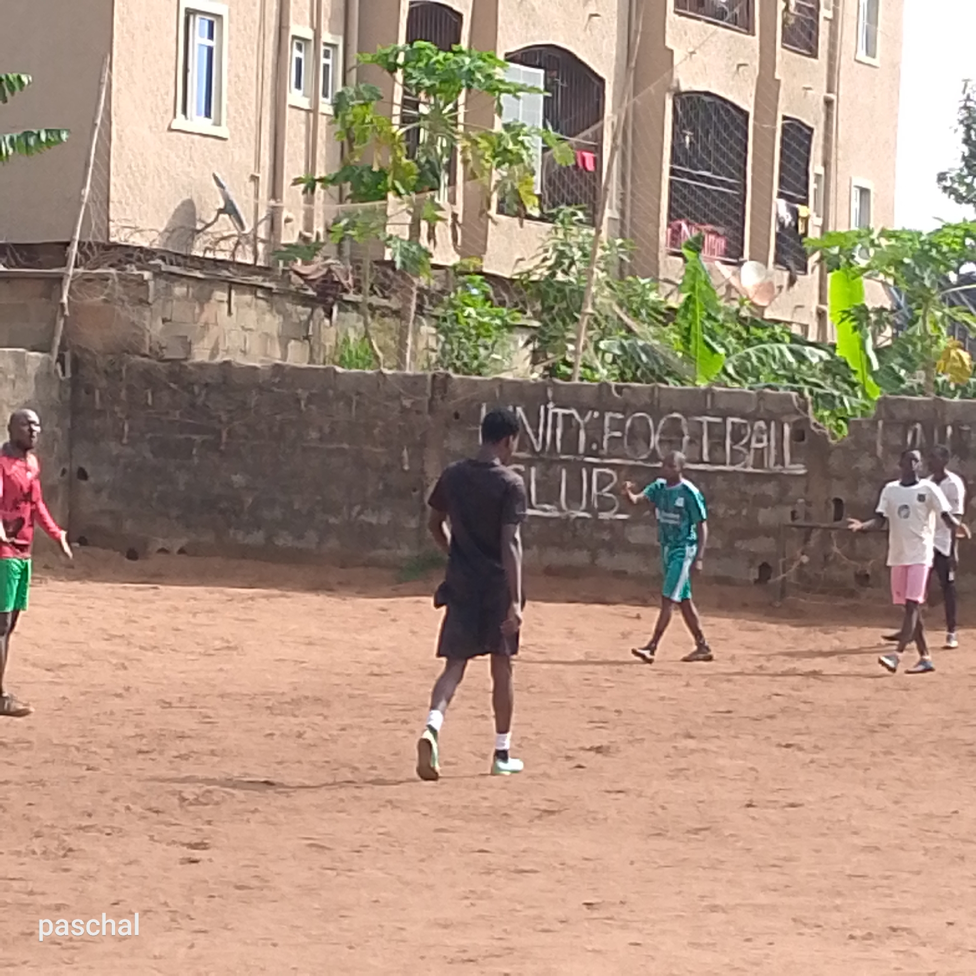 The image depicts a group of young men on a reddish-brown dirt field in Ukwu-Abwa, Nigeria. A partially visible building is present in the background, with a brick wall separating the field from the structure. Painted in white on the wall is "UNITY FOOTBALL CLUB". There are four young men are visible on the field. One man on the left wears a red long sleeve shirt and green shorts. In the center, a man with his back towards the viewer is wearing a dark short sleeve shirt and dark shorts with white socks and shoes. To his right, a boy in a teal uniform is seen. And to the right of him, two boys are seen in light color shirts and shorts. The ground appears dusty. The sun appears to be brightly out.