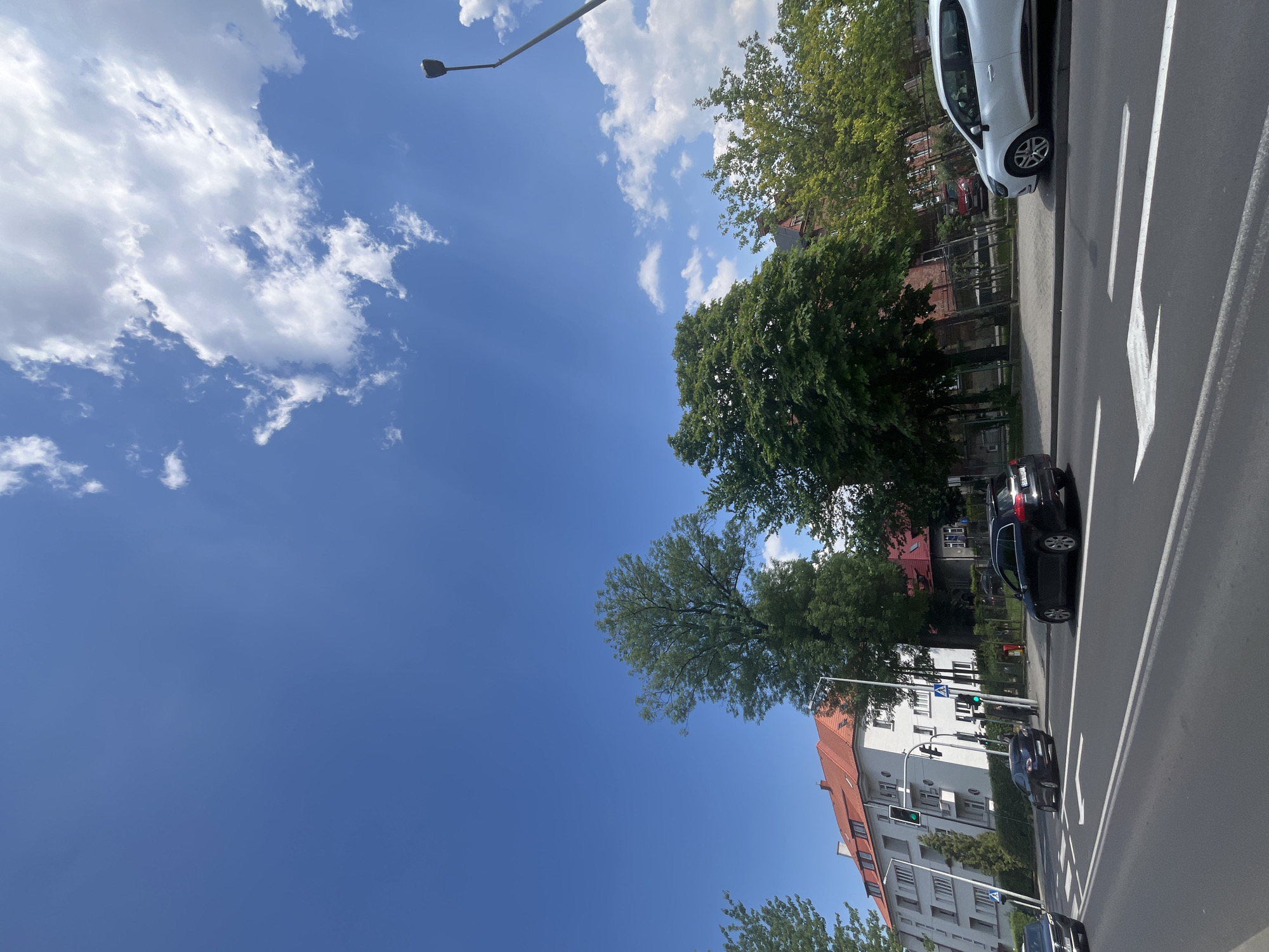 The image captures an outdoor urban or suburban street scene on a bright, sunny day. The sky is a vivid blue, adorned with scattered white cumulus clouds, and faint sun rays are visible in the upper left portion of the frame.
A wide asphalt road occupies the right side of the image, featuring distinct white road markings including solid lines, dashed lines, and prominent arrows indicating turn directions. Several vehicles are present: a white hatchback is parked along the roadside in the foreground, followed by a dark-colored sedan or hatchback. Further down the street, two more dark cars are visible, with the lead car stopped at a traffic light that displays a green signal, suggesting active traffic flow. The license plate of the dark car in the mid-ground begins with "W", which is a characteristic identifier for Polish vehicle registration plates, particularly from the Warsaw region.
Lining the street and situated behind the buildings are numerous lush green trees, contributing to a verdant urban landscape. On the left side of the road, a multi-story apartment building with a light-colored facade and a distinctive reddish-brown tiled roof stands prominently. It features multiple windows and visible balconies. Other residential or commercial buildings with similar architectural styles and reddish roofs are partially glimpsed through the trees in the background. A tall street lamp extends upwards from the left, and another traffic light pole is visible further down the street near the apartment building.
The scene conveys a sense of everyday urban activity under clear, fair weather conditions during daylight hours. No people are discernible in the image.