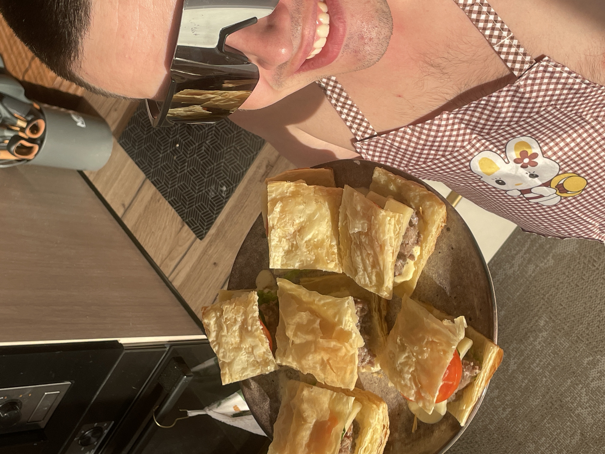 A man wearing mirrored sunglasses and a brown and white checkered apron with a cartoon bunny design is holding a plate of square pastries with visible fillings. The pastries appear to contain meat, tomato, and cheese. The setting appears to be indoors, possibly a kitchen, given the presence of a stove and patterned countertop. The specific location of Подшивалово, Russia is not definitively discernible from the image.