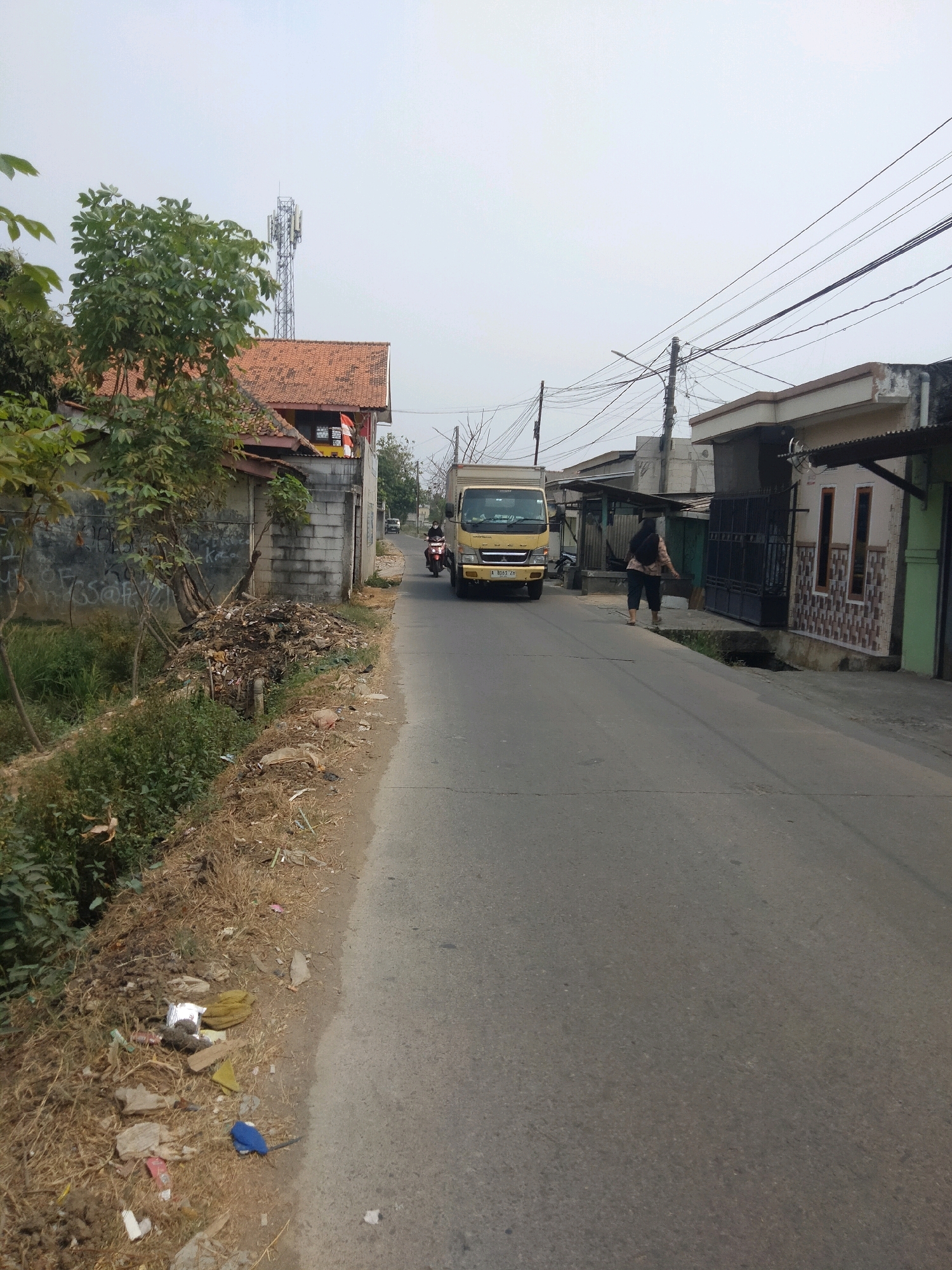 The image depicts a street scene in Suka Sari, Indonesia. A light-colored, overcast sky dominates the upper portion of the frame. On the left side of the road, a pile of trash is visible along the grassy verge, adjacent to a concrete wall with graffiti. A tree with green leaves grows near the wall. A red-tiled roof house is partially visible behind the tree and wall. Further down the street, a yellow truck with its license plate "A 8165 ZH" is driving away from the camera. A person on a motorcycle is approaching the truck. On the right side of the road, several buildings with simple facades and awnings line the street, along with power lines and utility poles overhead. A woman is walking away from the camera on the right sidewalk, which has a metal gate and decorative tilework on the buildings. The road itself is asphalt with visible cracks and markings.