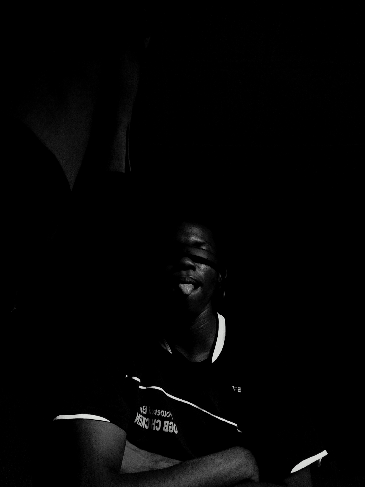 A black and white photo features a dark-skinned person in a dark environment. A strip of material covers their eyes. Their tongue is sticking out, and they are wearing a dark shirt with white trim and lettering (OGB CH CHEW). Their arms are crossed. There is a dark, amorphous shape in the upper left. No location information related to Keffi, Nigeria, can be ascertained from the image.
