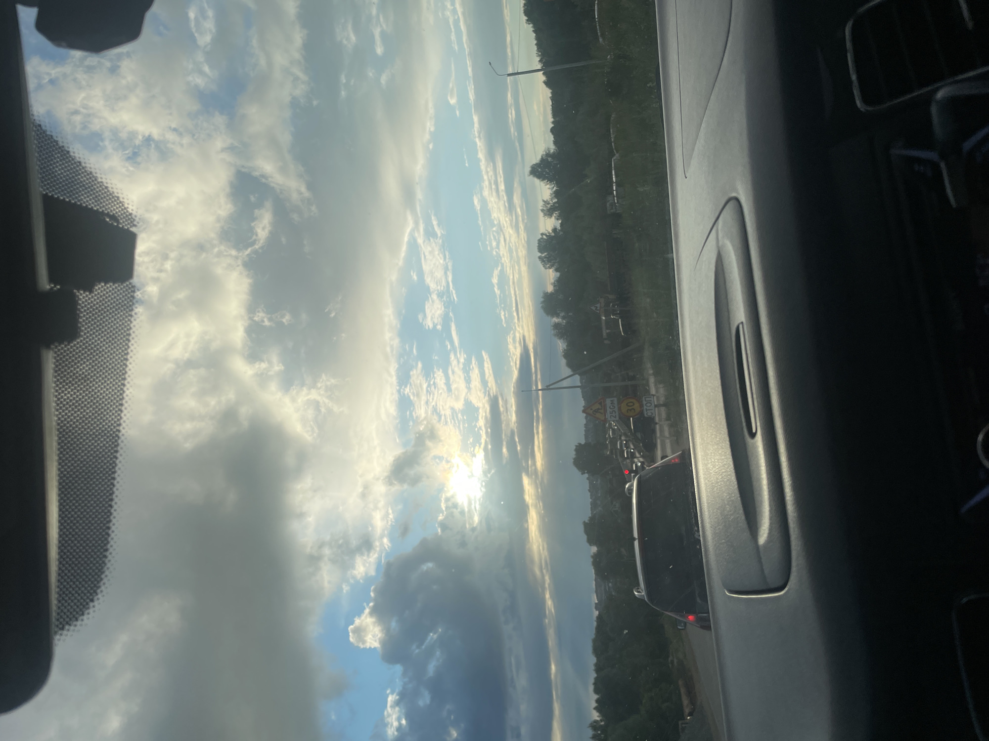 The image captures a scenic view from inside a vehicle, looking out through the windshield. The scene is dominated by a dramatic sky filled with a mixture of dark, heavy clouds and bright, sunlit patches, suggesting a dynamic weather pattern. The sun is partially obscured by the clouds, casting a warm, ethereal glow across the sky and illuminating the edges of the clouds with a golden hue.
In the distance, a treeline marks the edge of a landscape, likely rural or semi-rural, given the presence of vegetation. Several vehicles are visible on a road, indicating that the car the photo is taken from is in traffic or moving along a route. The presence of road signs suggests a developed area, though the overall impression is one of natural beauty contrasting with everyday infrastructure. One sign is partially visible, featuring a red circle with the number "30" inside, likely indicating a speed limit. Another sign says "СТОП," which means "STOP" in Russian.
The interior of the car is partially visible, with the dashboard and a portion of the passenger seat contributing to the frame. The perspective is from the driver's or front passenger's seat. The time of day appears to be late afternoon or early evening, given the angle of the sun and the quality of the light. The weather is clearly transitioning, with both cloudy and sunny elements present. The overall mood is one of quiet observation of the natural world from within the controlled environment of a vehicle, possibly during a commute or a journey. The location context of Gavrilovka, Russia, adds a specific geographical anchor to the scene.