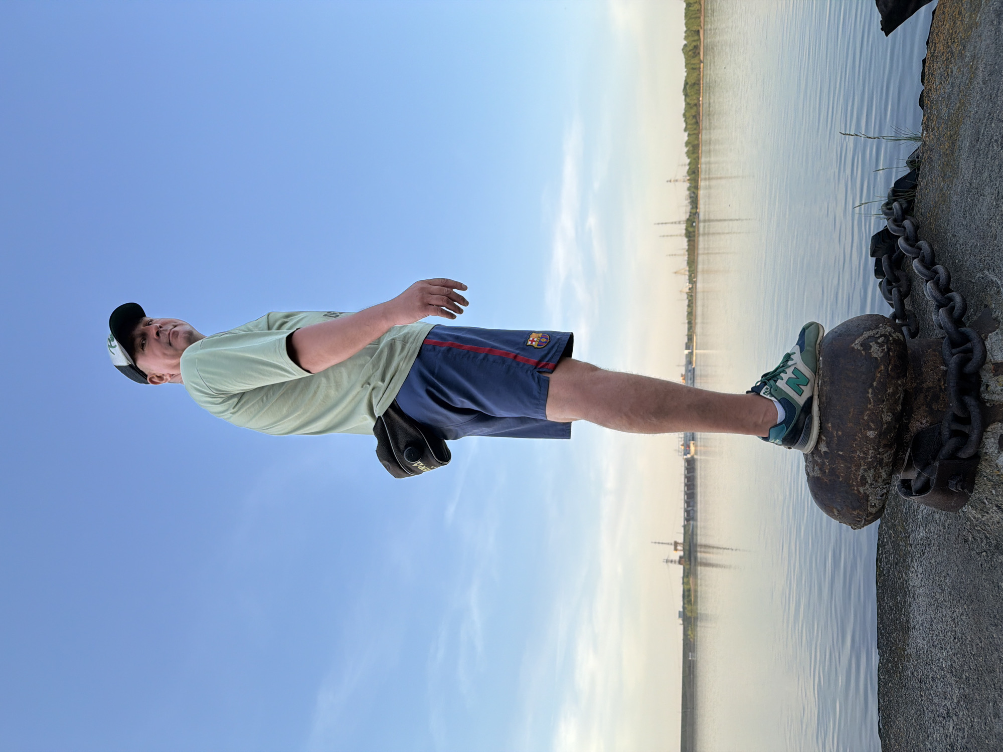 A man stands on a pier in Chaikovsky, Russia. He wears a light green shirt, navy blue shorts with a red stripe and a Barcelona FC crest, a black and white baseball cap, and green and white sneakers. He stands with one foot on a large, rusty metal bollard secured to the pier by heavy chains. The pier is made of gray concrete. In the background, a body of water reflects the sky. On the opposite shore, trees and structures, including possible industrial elements, are visible. The sky is a clear blue gradient.