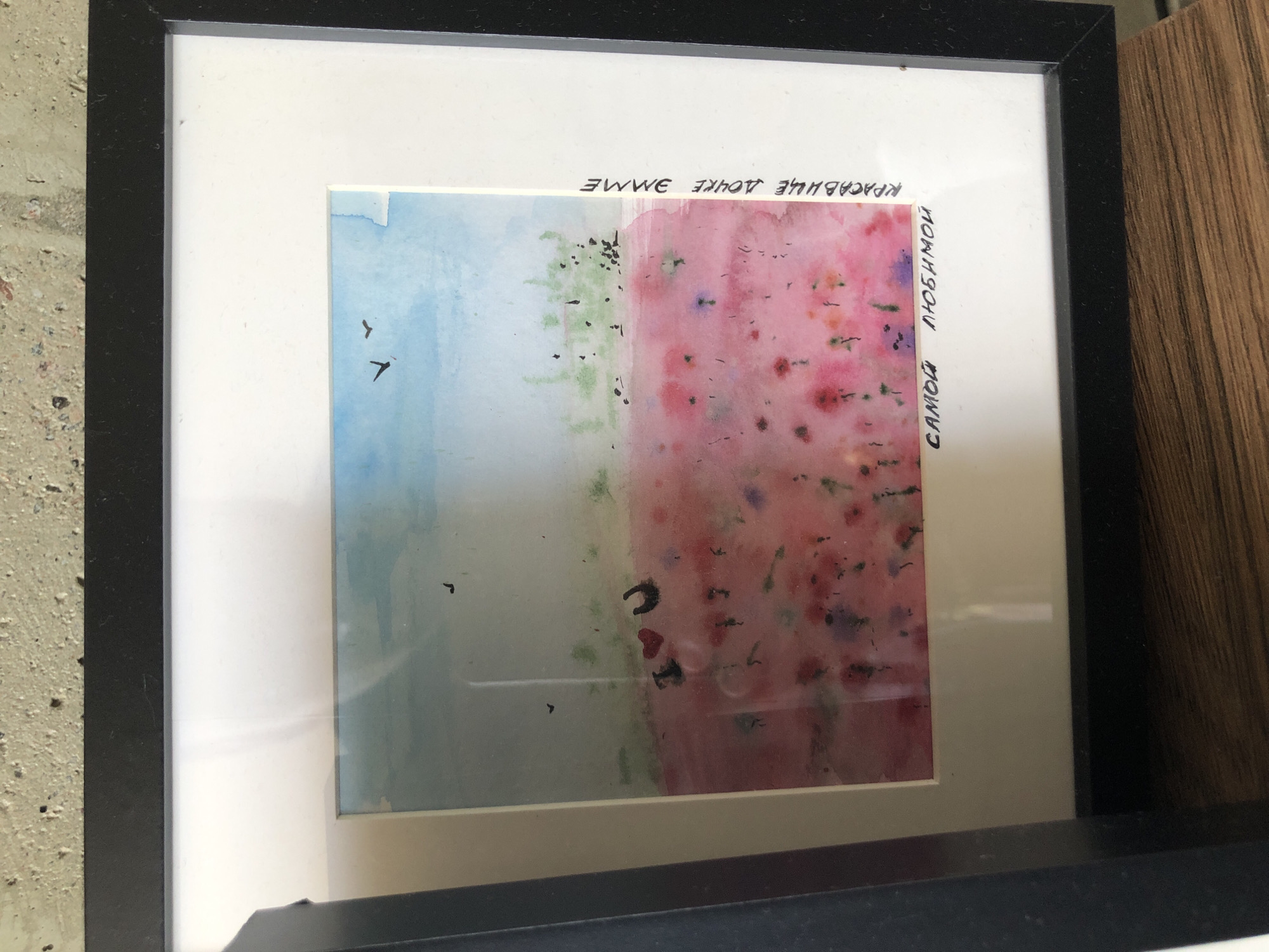 The image shows a framed watercolor painting. The painting depicts a field of pink and red flowers under a light blue sky with two birds flying. There are also some dark specks that could represent more flowers or foliage. On the left side of the painting, there are two red hearts and a black horseshoe shape. The painting is matted with a white border and framed in black. There is text written in black ink on the white mat. On the right side, vertically oriented, it reads "КРАСАВИЦЕ ДОЧКЕ ЭММЕ" which translates to "To beautiful daughter Emma." At the bottom, horizontally oriented, it reads "САМОЙ ЛЮБИМОЙ" which translates to "To my dearest." The painting is placed on a wooden surface, possibly a shelf or table, against a textured wall. There is no discernible location in Saint Petersburg visible in the image.