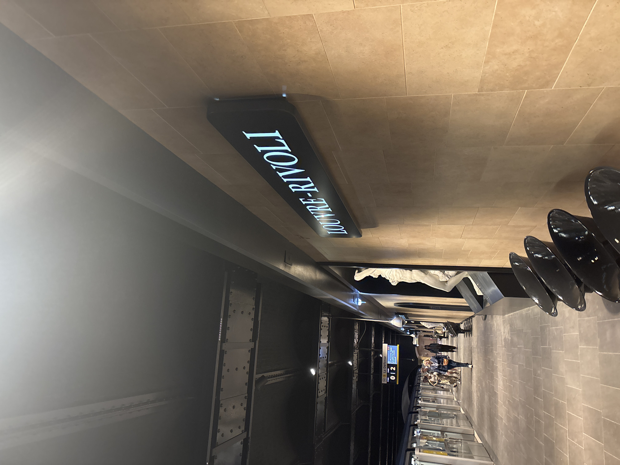 The image shows the Louvre-Rivoli metro station in Paris, France. An overhead sign displays "LOUVRE-RIVOLI" in large, illuminated lettering. People are walking in the station. There are metallic structural elements, and art objects are visible, including what appears to be a sculpture. Signage displays "La Defense" and "012". The overall environment suggests a subway station. The scene is lit with artificial lighting.