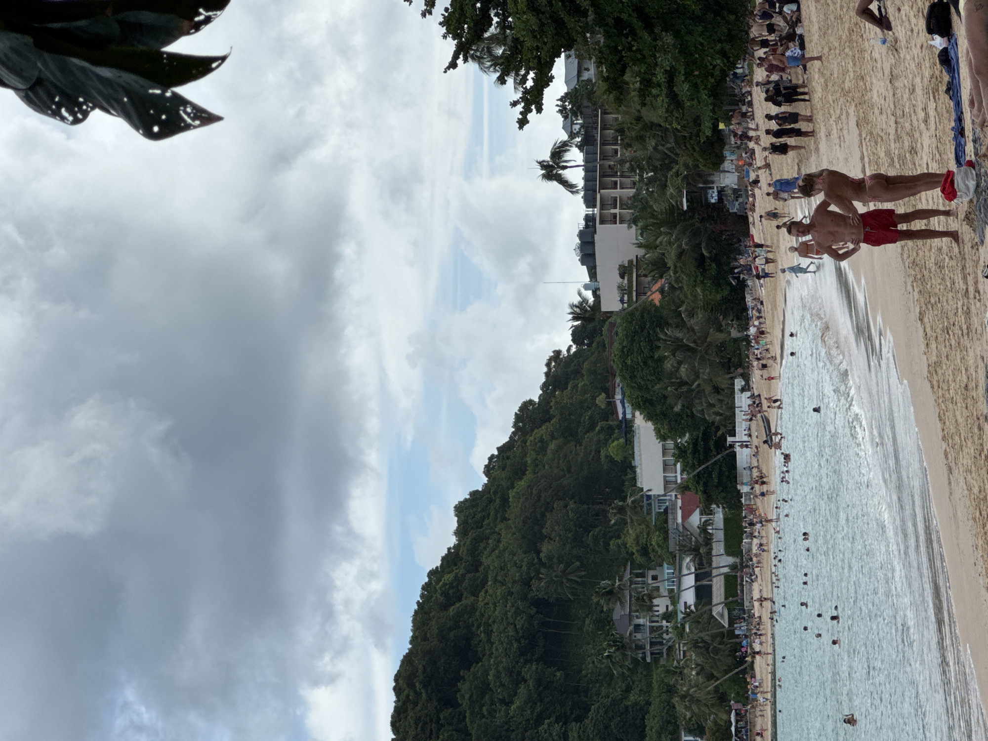 The image depicts a crowded beach scene on a cloudy day in Wichit, Thailand. The foreground shows a sandy beach with several individuals, including a couple in red shorts and a pink bikini standing and facing away from the camera. The water is turquoise and filled with numerous swimmers. In the background, a hillside covered in dense green trees slopes upwards, with several buildings and hotels interspersed among the foliage. Palm trees are visible throughout the landscape. The sky is predominantly overcast with large, grey clouds, but patches of blue sky are visible. The overall atmosphere appears to be one of a popular tourist destination during a somewhat dull weather day.