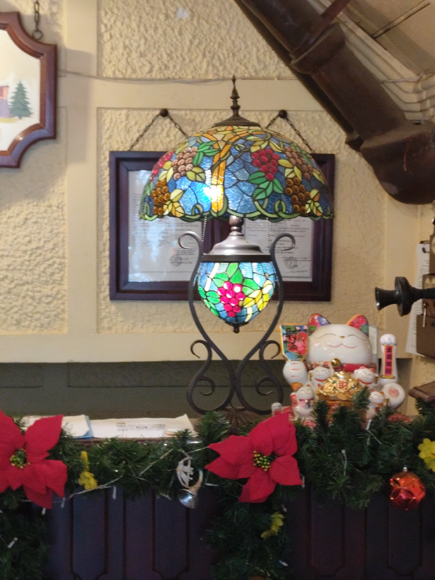 The image depicts a festive interior scene, likely a restaurant or shop, adorned with Christmas decorations. Dominating the center is a Tiffany-style stained glass lamp with a floral and grape motif, casting a warm glow. To the right of the lamp, a collection of "maneki-neko" (beckoning cat) figurines are arranged, some holding gold coins, signifying good fortune. The foreground features a lush garland of dark green pine branches, interspersed with vibrant red poinsettias and small, twinkling lights. A few yellow decorative elements and a red Christmas ornament are also visible on the garland. In the background, a framed artwork depicting a house and a Christmas tree hangs on a textured yellow wall, and another framed document is visible behind the lamp. A dark pipe runs diagonally across the upper portion of the image, suggesting an industrial or older architectural element. The overall impression is one of a cozy and welcoming atmosphere, characteristic of a holiday season in a place like Da Lat, Vietnam, which is known for its French colonial architecture and cooler climate that often lends itself to a festive Christmas spirit.