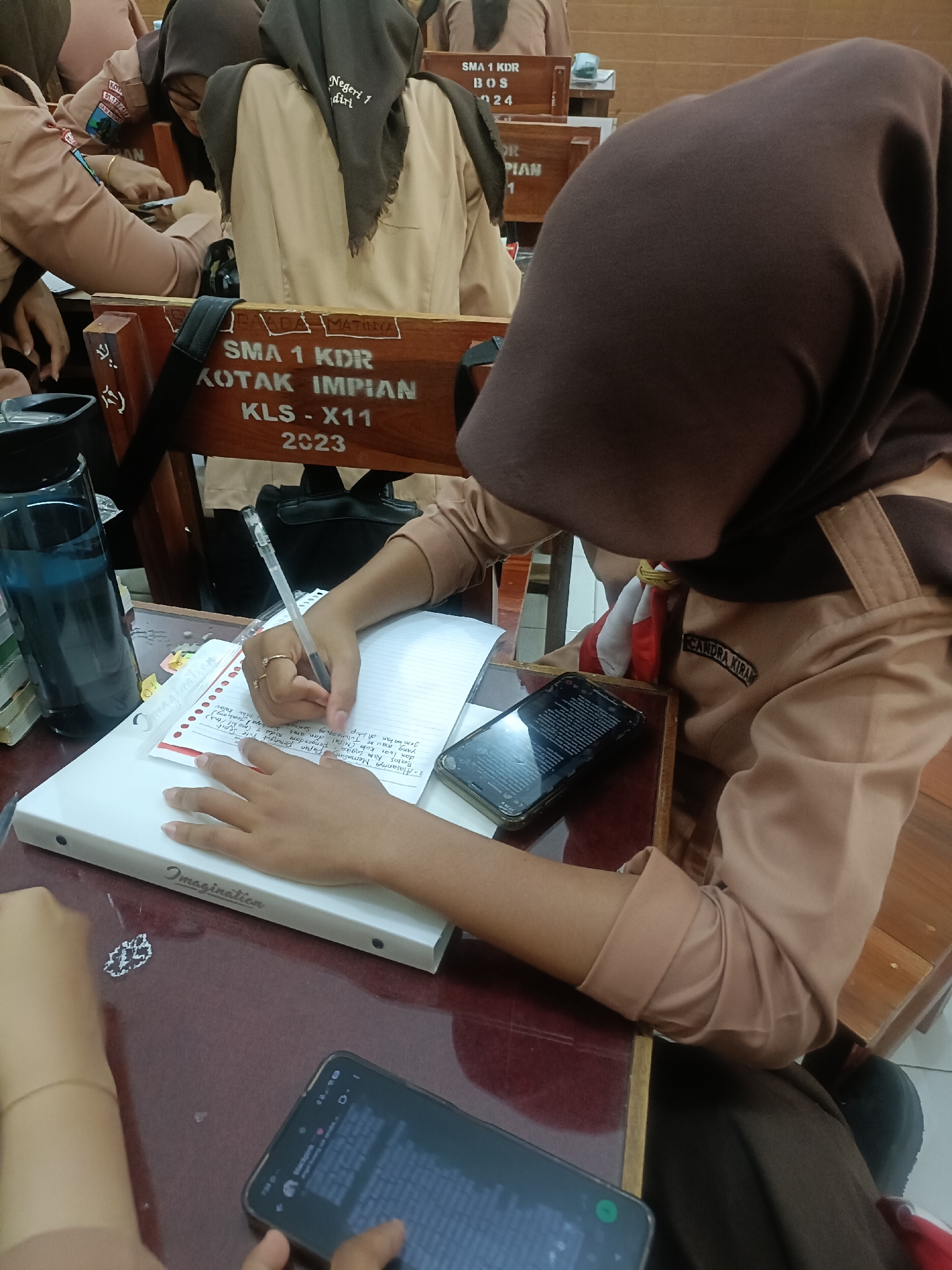 The image shows a classroom scene in Indonesia. The focus is on a student wearing a brown uniform and a headscarf, sitting at a desk and writing in a notebook. Another student is partially visible in the foreground, also wearing a similar uniform. The desk has various items on it, including a water bottle, a mobile phone, and a binder with the word "Imagination" printed on it. In the background, other students and classroom furniture are visible. Wooden chairs with text indicating "SMA 1 KDR" are also present, suggesting the location is a high school in Kediri City, Indonesia. The overall atmosphere appears to be one of a typical school day.