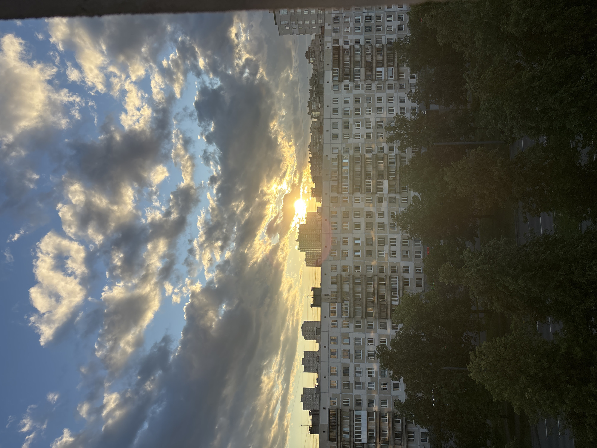 The image captures a city view at sunset, likely in Russia given the architectural style of the buildings. A large, multi-story apartment building dominates the mid-ground, its facade featuring numerous windows and balconies. In front of this building, a line of mature trees with dense green foliage partially obscures the lower levels. The sky above is filled with a dramatic array of clouds, ranging from dark, heavy masses to lighter, illuminated formations catching the golden rays of the setting sun. The sun itself is partially visible, peeking through the clouds and casting a bright glow. In the background, other buildings, including some taller structures, can be seen against the colorful sky. The foreground shows a glimpse of a street or road with a few visible lane markings, and more trees.