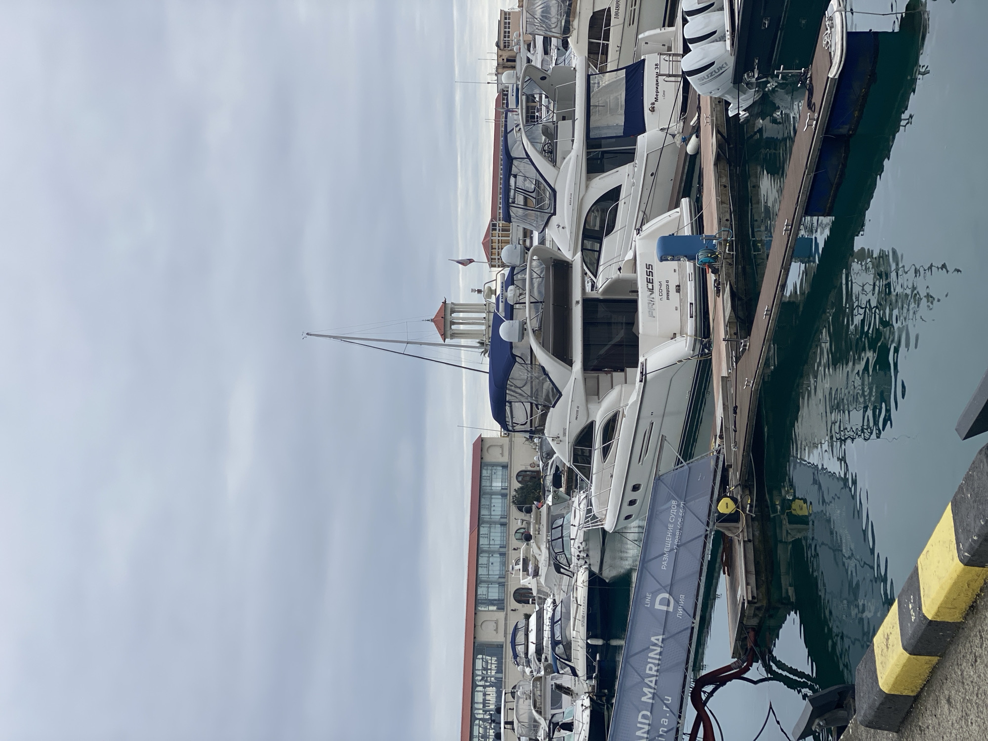 This image depicts the Grand Marina Sochi in Sochi, Russia, under an overcast sky. The scene is dominated by numerous recreational vessels, primarily white motor yachts and some sailboats, moored within the marina basin.
In the mid-ground, a white motor yacht with "PRINCESS п. СОЧИ ПРИНЦЕССА 45" (Princess Port Sochi Princess 45) is clearly visible on its stern. Adjacent to it is another yacht marked "Меридиан 38" (Meridian 38). Many vessels feature blue canvas bimini tops or covers, and several are equipped with radar domes, some bearing the "FURUNO" brand. Outboard engines displaying "SUZUKI" are visible on a boat in the lower right.
The vessels are moored to dark-colored floating docks. A blue service pedestal, likely for power and water, is present on one of these docks near the "Princess" yacht. In the immediate foreground, a black and yellow striped concrete barrier marks the edge of the quayside. A large, grey-blue banner or ramp cover extends across the lower left portion of the frame, partially obscuring the water, displaying text in English and Russian, including "LAND MARINA," "ЛИНИЯ D" (Line D), and "РАЗМЕЩЕНИЕ СУДОВ" (Vessel Mooring) along with a phone number.
The background features architectural elements: a large building with a terracotta-tiled roof and a facade of numerous arched windows, typical of waterfront development. To the right of this building, a distinctive white, classical-style rotunda with a red conical roof is visible. A Russian flag flies from a flagpole located near this rotunda. The water in the marina is calm, reflecting the boats and the overcast sky with a dark teal hue. No individuals are visible in the scene.