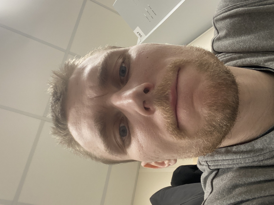 A direct, low-angle headshot captures a light-skinned adult male, presumably located in Nikol'skoe, Russia. The male has blue eyes and is looking directly at the camera. His short, light brown hair is disheveled on top. A blonde-to-light brown mustache and a short beard cover his upper lip, chin, and jawline. His forehead displays several horizontal lines. He is wearing a grey, possibly hooded, garment, visible around his neck and shoulders. The background consists of a light-colored dropped ceiling with a grid of square or rectangular acoustic panels. To the subject's left, a dark, indistinct fabric or object is partially visible. To his right, a light-colored vertical surface, possibly a cabinet or partition, displays the black numbers "567" and some smaller, illegible text. The scene is illuminated by overhead artificial lighting.