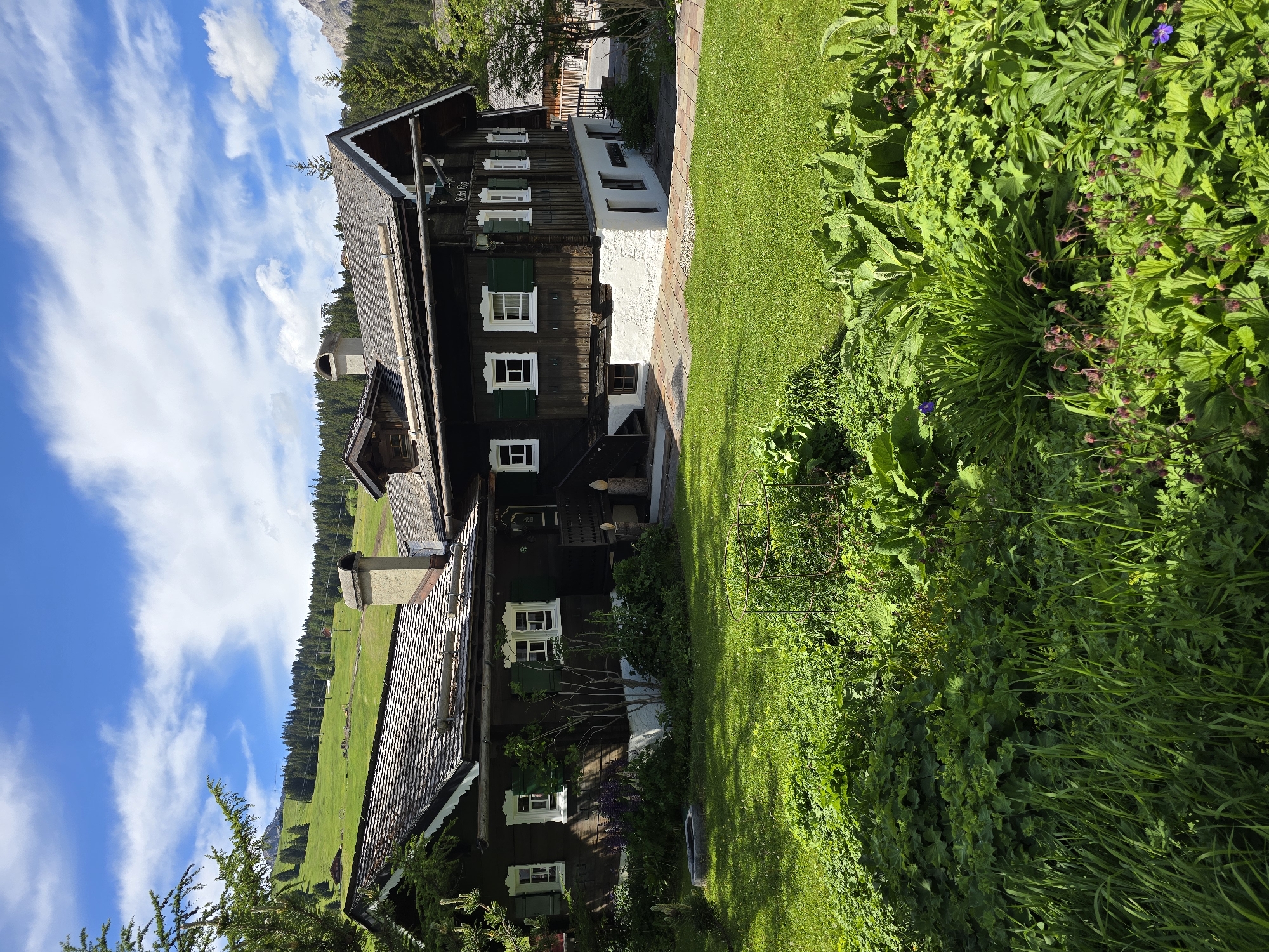 The image shows a traditional wooden house with a shingled roof, dark brown wooden siding, and green shutters on the windows. The house is situated in a lush green landscape with rolling hills, pine trees, and a bright blue sky with white clouds. In the foreground, there is a manicured lawn with various plants and flowers. The house has a sign that reads "Gästehaus." The setting suggests it is likely a mountain resort area, possibly in the Austrian Alps given the architectural style and landscape. The scene is tranquil and picturesque, evoking a sense of a peaceful retreat.