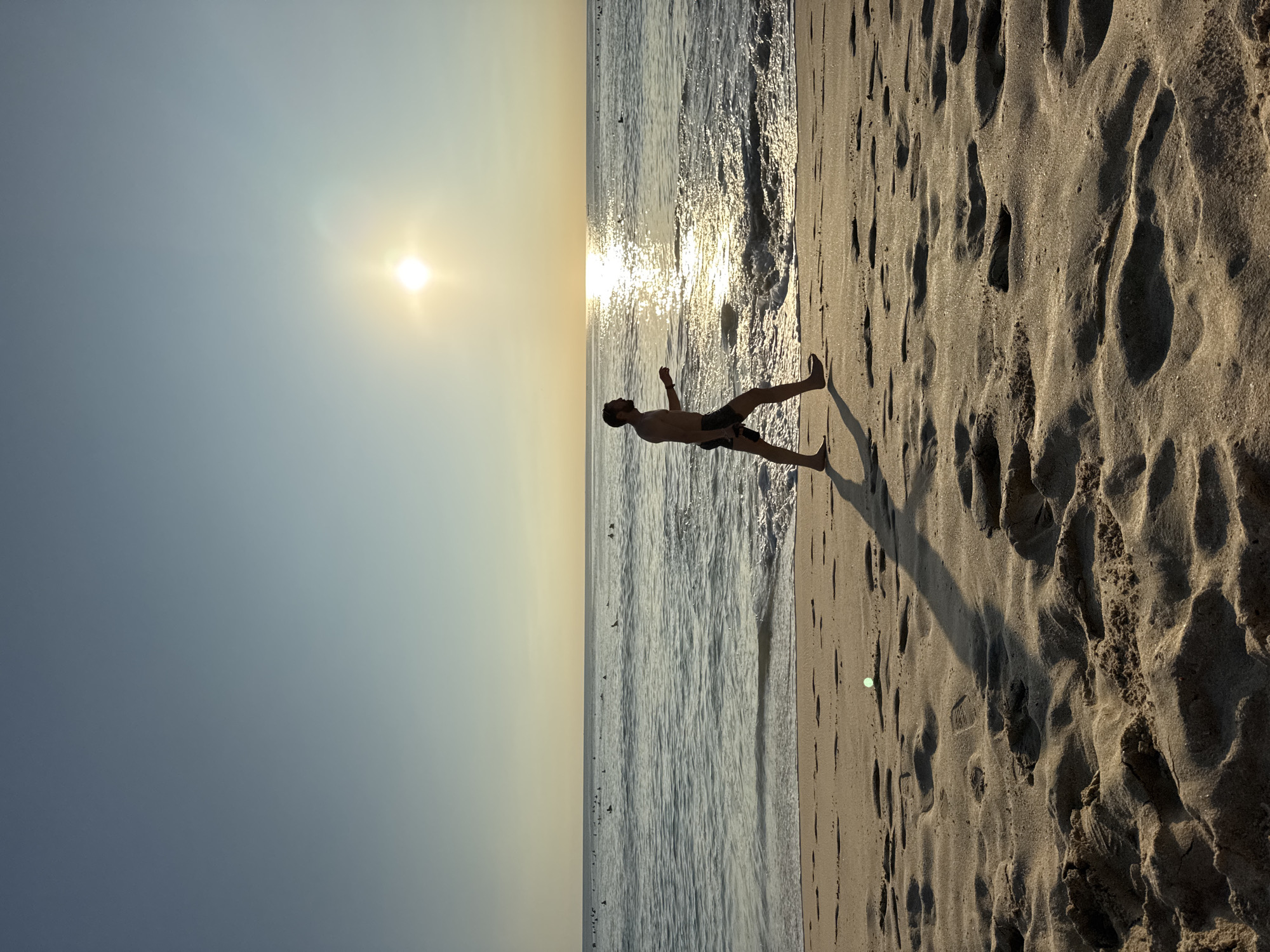A medium, tilted shot depicts a man standing on a beach with the ocean to his left and sand to his right. The sun is positioned high in the sky, casting a long shadow from the man onto the sand. The scene appears to be Kuta Selatan, Indonesia, based on the coastal landscape and sandy terrain. The man is facing towards the ocean, and his silhouette is visible against the bright light reflecting off the water. Waves are lapping the shoreline. The sand shows numerous footprints.