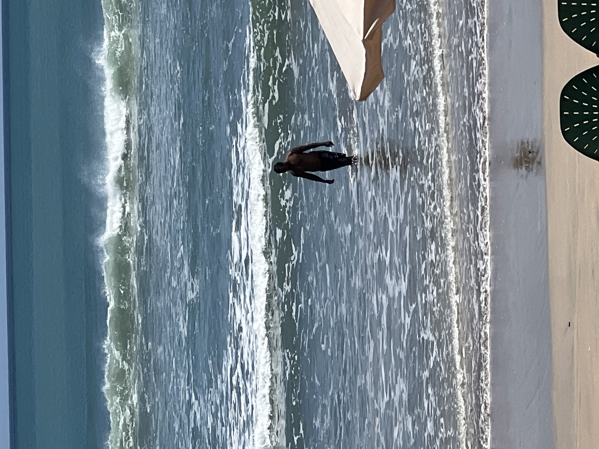 A man walks in the shallow water of a beach in Kuta, Indonesia. The man is dark-skinned and appears to be wearing dark shorts. He is facing towards the right of the frame. The water is a dark blue and has waves breaking in the background. The sky is a clear blue. In the foreground, there is a beach umbrella and two empty chairs.