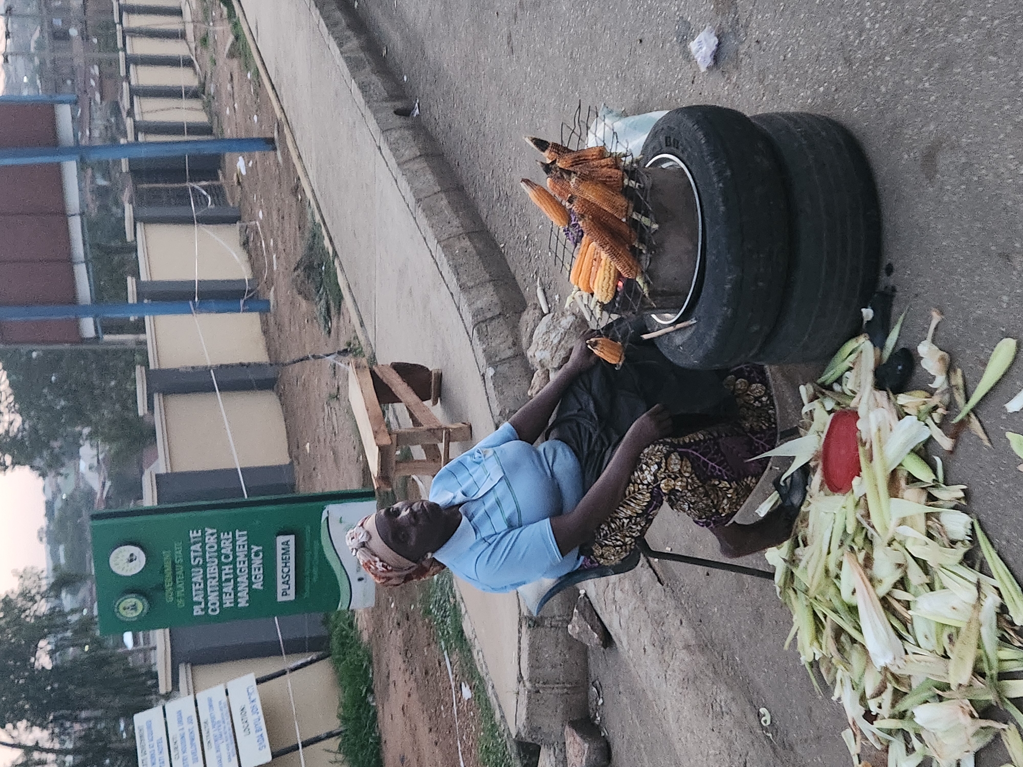The image shows a woman in Jos, Nigeria, selling roasted corn at a roadside stand. The stand is built on top of tires, with a grill holding the corn over burning coals. The woman is sitting on a stool next to the grill. The background features a building with a sign indicating the "Plateau State Contributory Health Care Management Agency (PLASCHEMA)." There are corn husks lying on the ground. The location is specified as Sada Biyu, Jos.