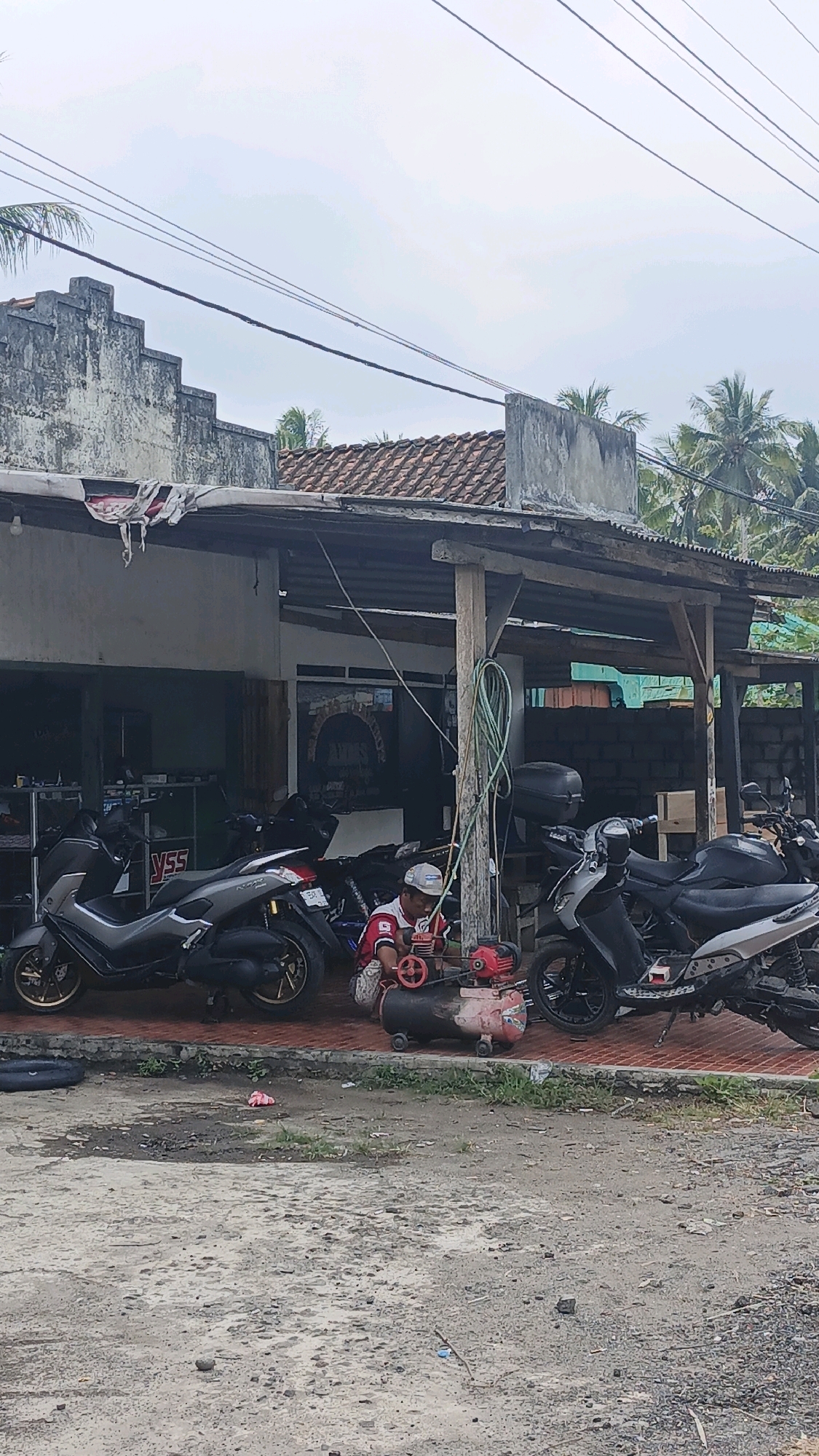 In Cibenda, Indonesia, a person in a red and white shirt and a white cap is working on a red air compressor next to several motorcycles parked on a tiled surface in front of a building. The building has a corrugated metal roof and a weathered facade, with a covered area extending outwards. The ground in front of the building is rough and appears to be a dirt or gravel lot. Several power lines are visible overhead, against a cloudy sky. Palm trees can be seen in the background.