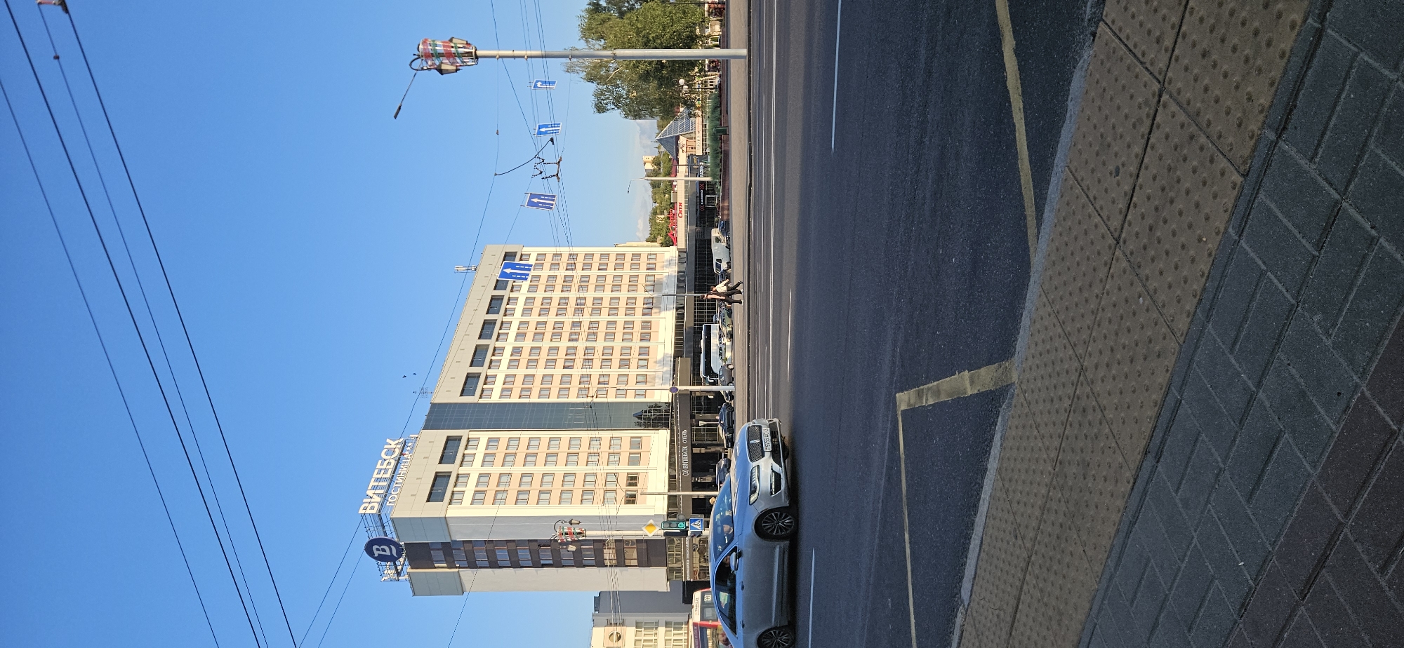 The image captures a view of the "VITEBSK" hotel, a prominent building in the city of Vitebsk, Belarus. The hotel is a multi-story structure with a light-colored facade and numerous windows. Above the entrance, a large sign displays the hotel's name in blue letters, along with the word "ГОСТИНИЦА" (hotel). The hotel is situated on a city street with vehicular traffic and pedestrians. A silver car is visible in the foreground, driving on the road. In the background, other buildings and trees can be seen. The sky is clear and blue, indicating a sunny day. The scene appears to be a typical urban setting with a mix of commercial and residential elements.