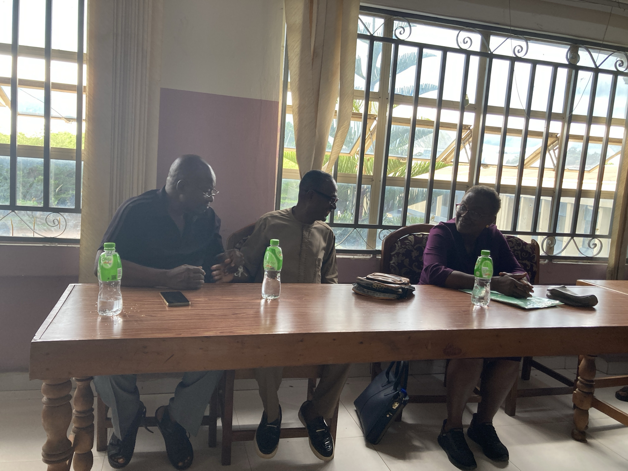 Three adults, two men and one woman, are seated around a long, polished wooden table in an indoor setting, likely a meeting room or office in Umuahia, Nigeria. Bright natural light streams in from a large window in the background, which features metal security bars with decorative scrollwork and light-colored pleated curtains.
On the left, an older man wearing a dark short-sleeved shirt and glasses is seated, with a green-capped "CHIVITA WATER" bottle and a black smartphone on the table in front of him. Next to him, a second older man in a light brown traditional African top (possibly a dashiki or kaftan) and glasses sits, looking towards the woman. His own "CHIVITA WATER" bottle and a small patterned clutch bag are on the table in front of him. On the right, an older woman in a dark purple long-sleeved top and glasses is seated, looking towards the middle man with a slight smile. A green folder or stack of papers and another "CHIVITA WATER" bottle are on the table in front of her, and a dark tote bag rests on the floor beside her.
The individuals appear to be engaged in a calm and focused conversation or meeting. The room has light-colored tiled flooring and white walls, with a section of the wall visible behind the middle man painted a light purple or pink hue. Outside the window, green foliage and what appears to be clear sky are visible, indicating bright daytime weather.