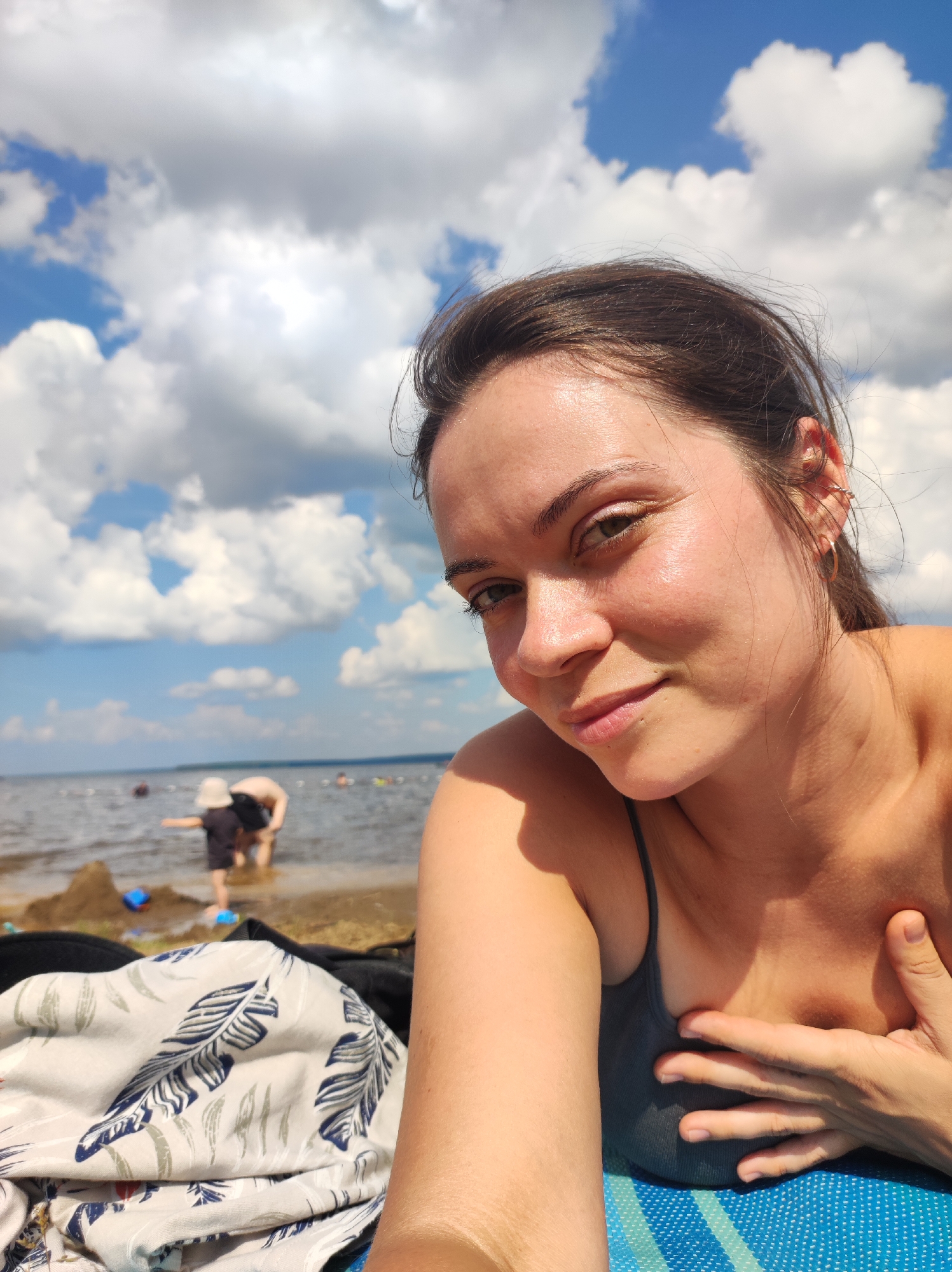 A woman is taking a selfie on a sunny beach in Chaikovsky, Russia. The sky is filled with white, puffy clouds. In the background, the Kama River is visible, with several people swimming and playing in the water. Two children are building a sandcastle near the water's edge. The woman is wearing a gray tank top and has her hand placed on her chest. A patterned beach towel is visible in the foreground.