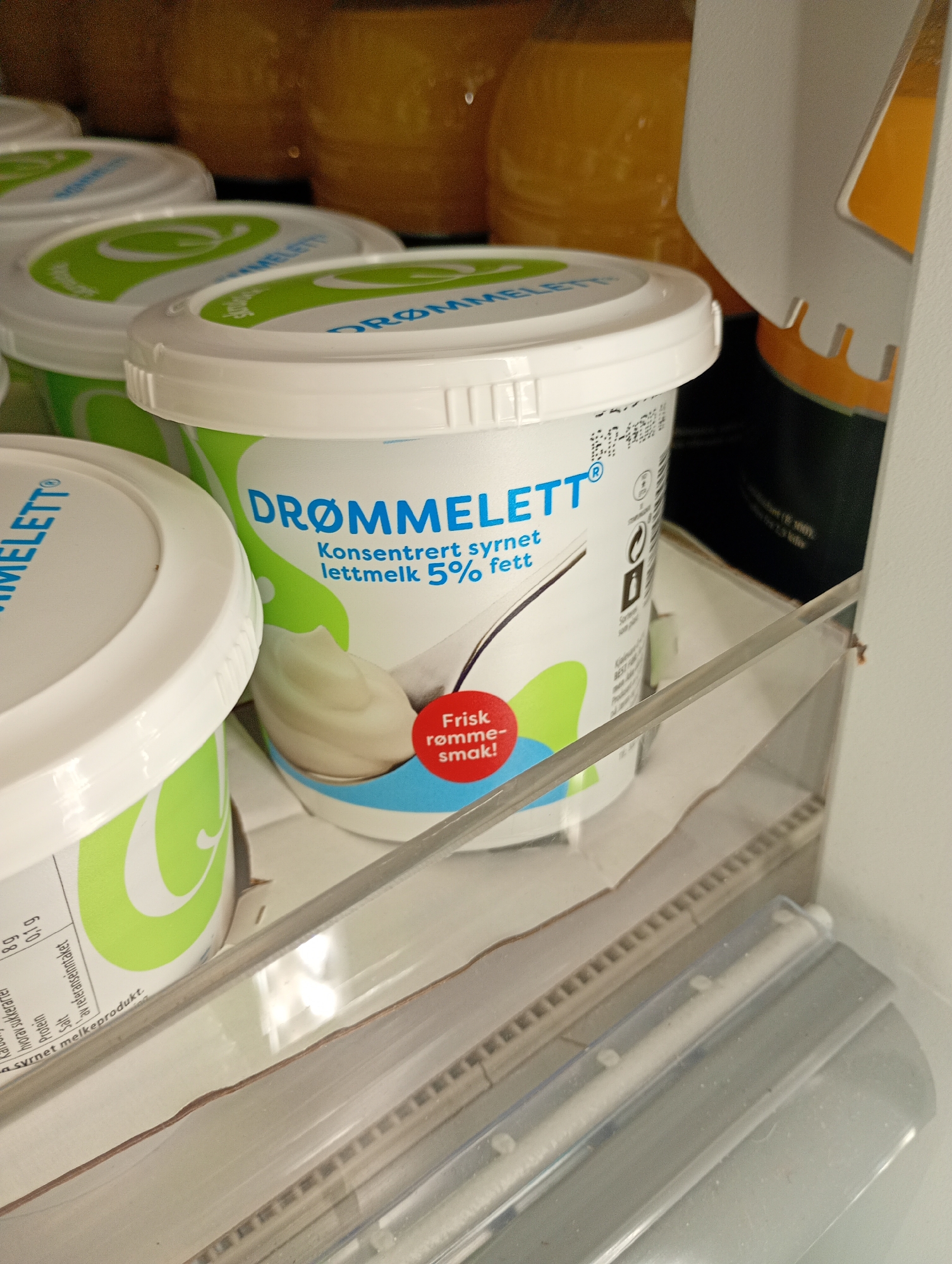 The image shows a close-up view of several containers of "DRØMMELETT" sour cream, a dairy product. The containers are white with green and blue branding. One container in the foreground is partially visible, showing the brand name and text that reads "Konsentrert syrnet lettmelk 5% fett," indicating it is concentrated sour milk with 5% fat. A red circular label on this container reads "Frisk rømme-smak!" (Fresh sour cream taste!). The containers are arranged on a shelf, possibly in a refrigerator, with other food items and packaging visible in the background and foreground. The lighting suggests an indoor environment.