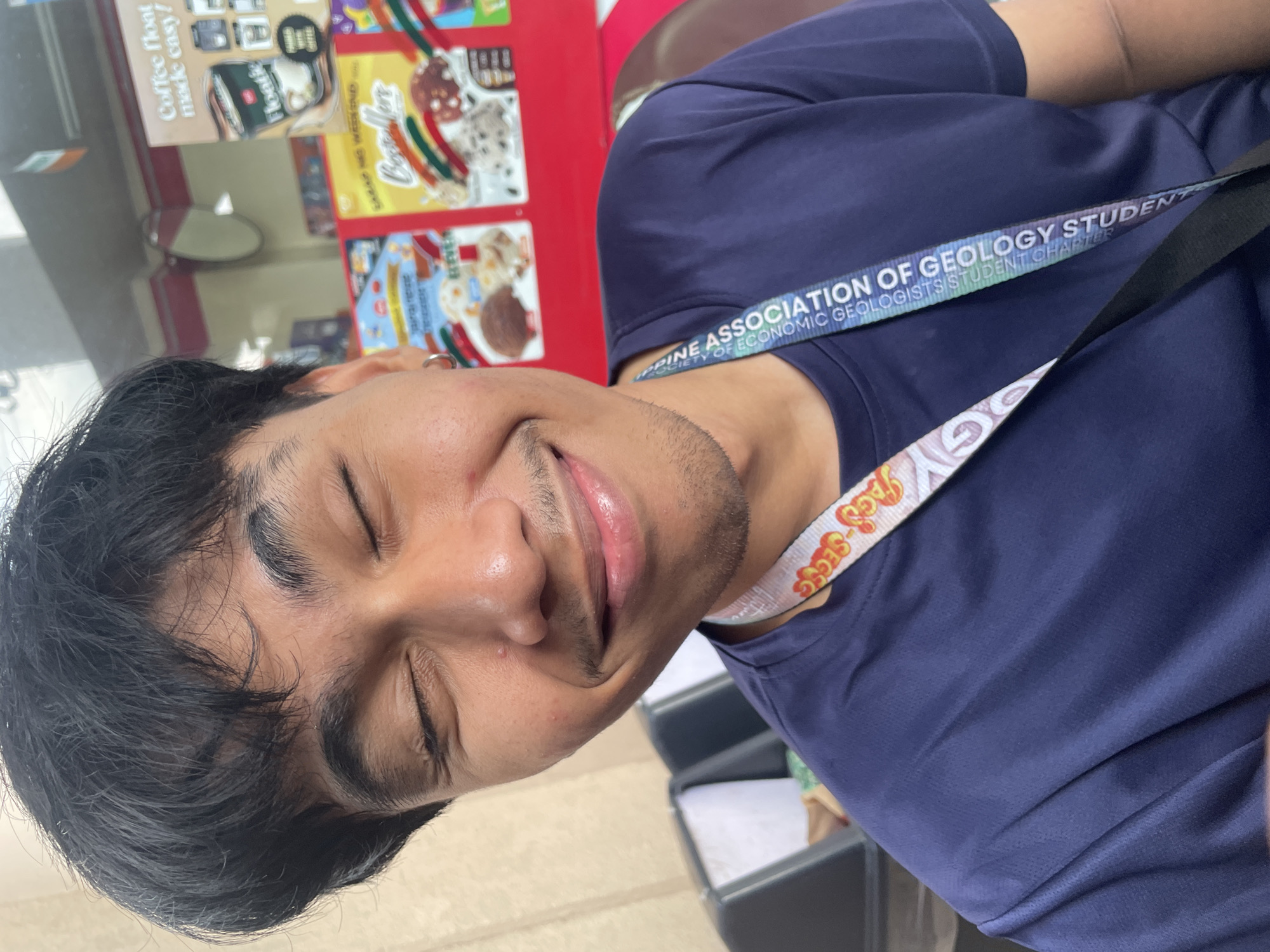 In a close-up shot, a young Filipino male with dark hair and a slight mustache is pictured with his eyes closed, smiling. He wears a navy blue t-shirt with a lanyard displaying text that includes "ASSOCIATION OF GEOLOGY STUDENTS" and "DAVAO CITY." The background suggests an outdoor setting, possibly near a convenience store or food stall, with posters for ice cream and coffee visible. The lighting is natural and bright. The overall impression is candid and informal.