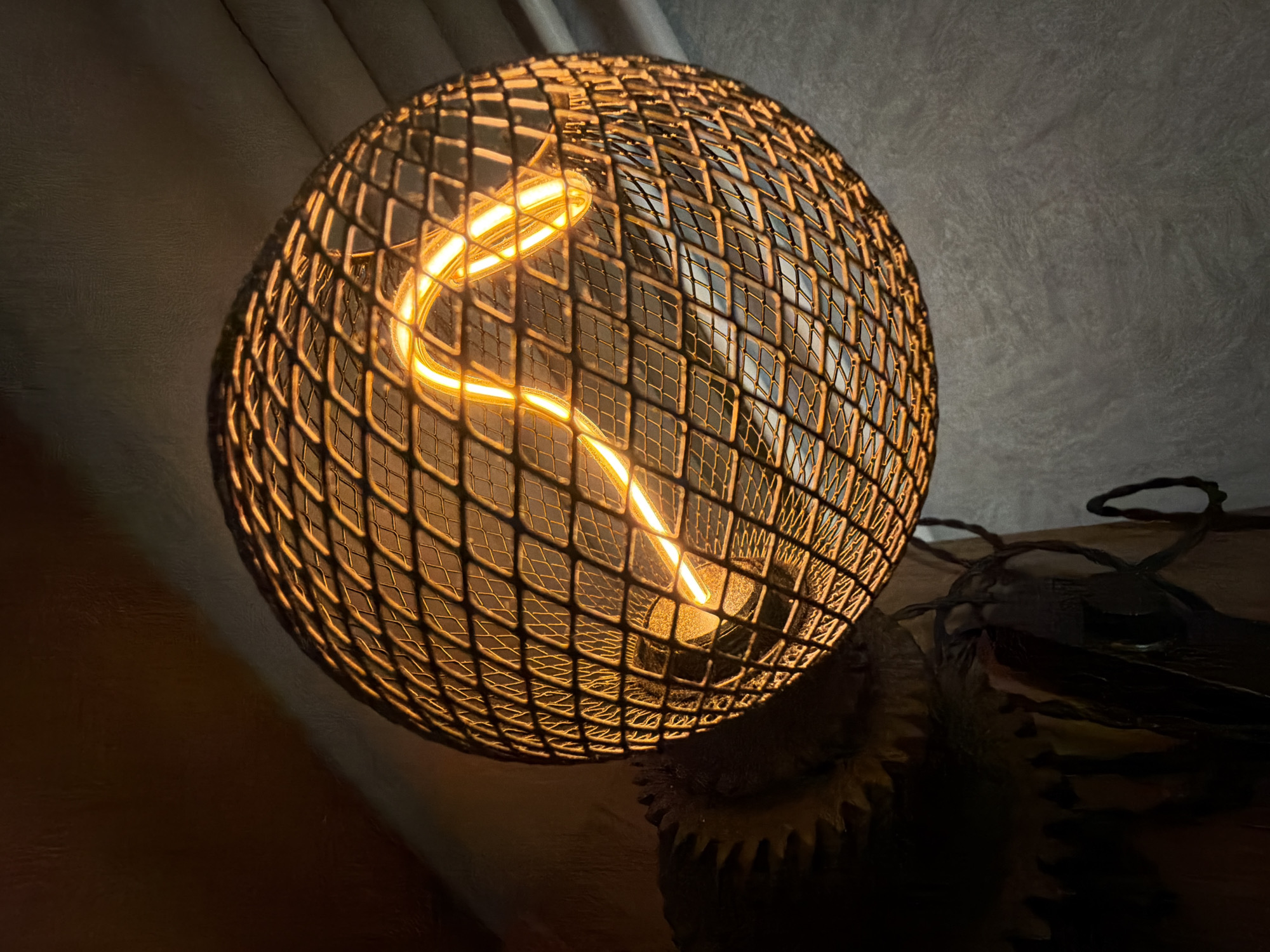 The image displays a close-up of a decorative lamp, creating a warm and inviting ambiance in a dimly lit indoor setting in Sofia, Bulgaria. The central subject is a spherical light fixture, characterized by a dark, metallic mesh cage that serves as a lampshade. Inside this cage, an Edison-style LED light bulb is visible, featuring a prominent, glowing, S-shaped filament that emits a soft, amber light.
The base of the lamp, partially visible towards the bottom right, appears to incorporate dark, gear-like or cogwheel elements, suggesting an industrial or possibly steampunk aesthetic. To the left and top, a muted, textured background, possibly a fabric curtain or wall, is bathed in the warm glow cast by the lamp. Below the lamp, a dark surface, likely wood, is visible, with dark cables or wires trailing off to the right side of the frame.
The overall scene is dark except for the light emanating from the bulb, indicating it is likely evening or night, or that the room is intentionally lit for a cozy atmosphere. No people or specific activities are depicted, and no visible text is present in the image. The focus is entirely on the illuminated lamp and its immediate surroundings.