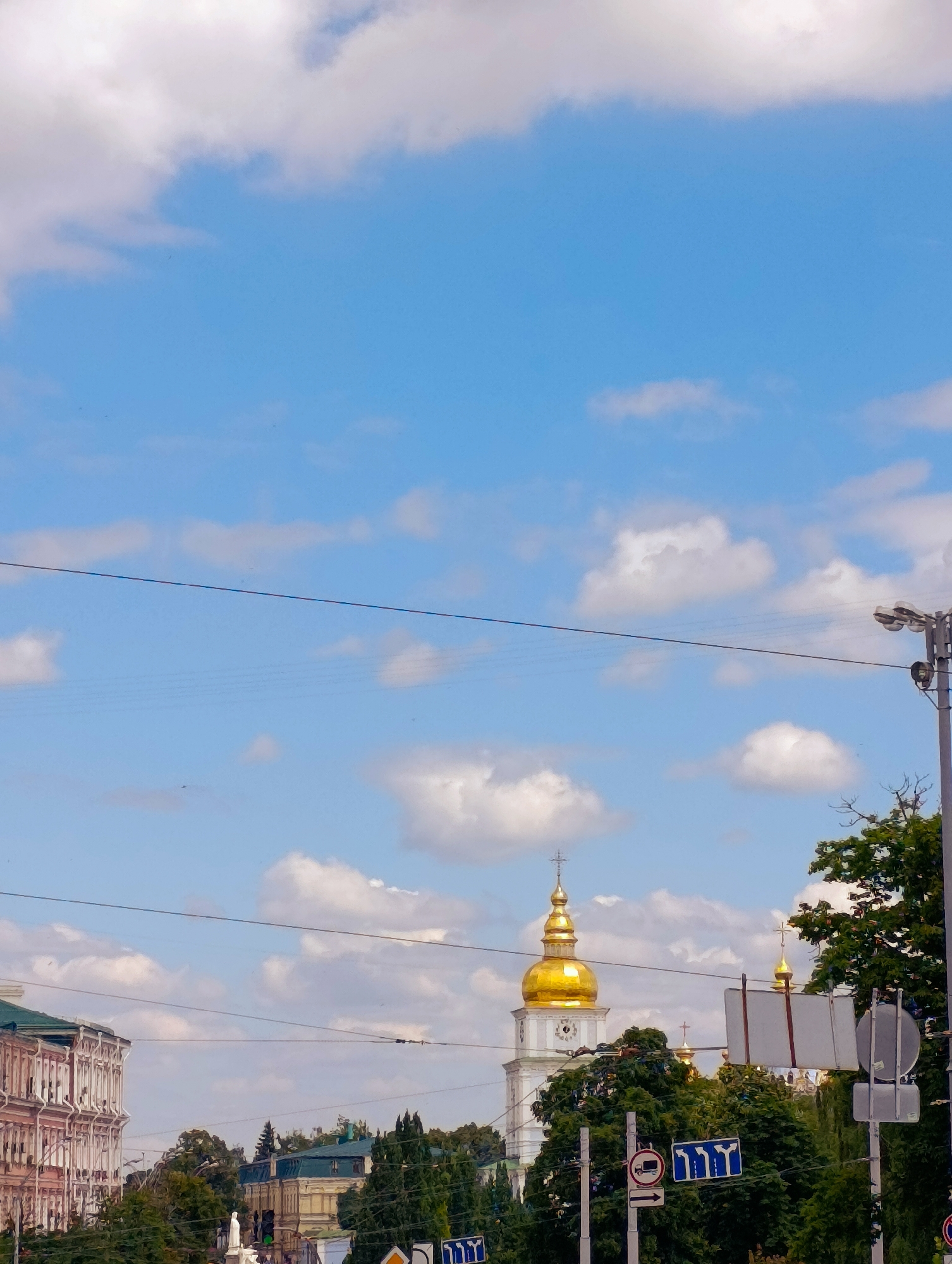 The image shows a view of Kyiv, Ukraine, with the iconic golden dome of the Kyiv Pechersk Lavra visible in the distance. The sky is a bright blue with scattered white clouds. In the foreground, there are power lines stretching across the frame. A street light pole with multiple lamps is on the right side of the image. Several trees line the middle ground, with their green foliage contrasting against the buildings and sky. On the left, a multi-story building with ornate architectural details is visible. Traffic signs, including a "no trucks" sign and a directional sign with arrows, are also present. The overall scene suggests a daytime view of an urban environment with historical and religious landmarks.