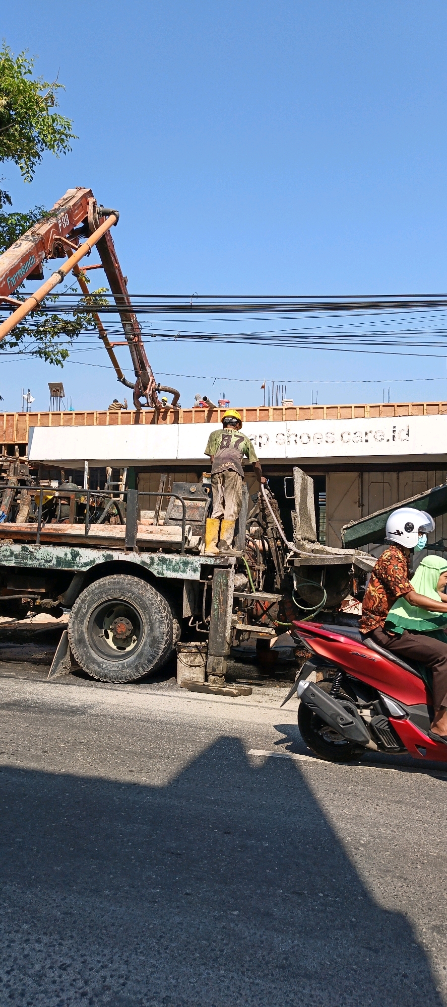 In Klaten, Indonesia, a construction site is active under a clear blue sky. A concrete pump truck, orange and bearing the markings "F33" and "Farrasingdo," is positioned to pour concrete onto the second story of a building under construction. Two construction workers, one wearing a yellow hard hat and beige work pants, stand on the truck. A second worker, also in a yellow hard hat and work pants, is partially visible on the roof of the building. The building's facade is white, with the text "ap shoes care.id" visible. In the foreground, a red scooter carries two individuals. The driver wears a patterned shirt and a white helmet with a blue mask, while the passenger wears a green hijab. The scene depicts a typical urban construction activity, with vehicles and people interacting within the environment.