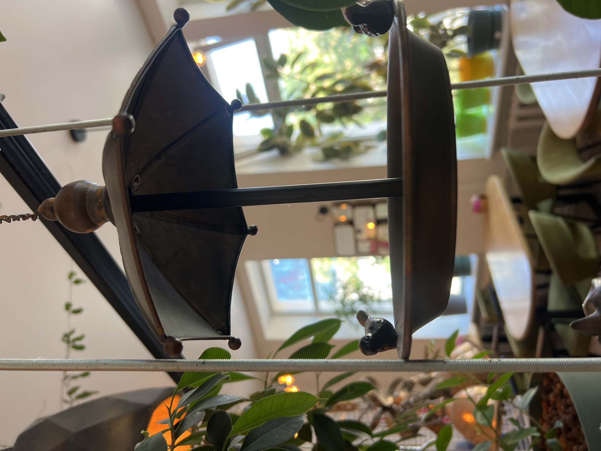 The image shows a close-up of a decorative bird feeder, shaped like a small umbrella, suspended indoors. The bird feeder is made of dark metal and has two small bird figurines perched on its rim. In the background, through a window, a sunlit scene with greenery and tables and chairs suggests it is a cafe or restaurant. The overall ambiance is cozy and decorated with plants. The location is Odesa, Ukraine.