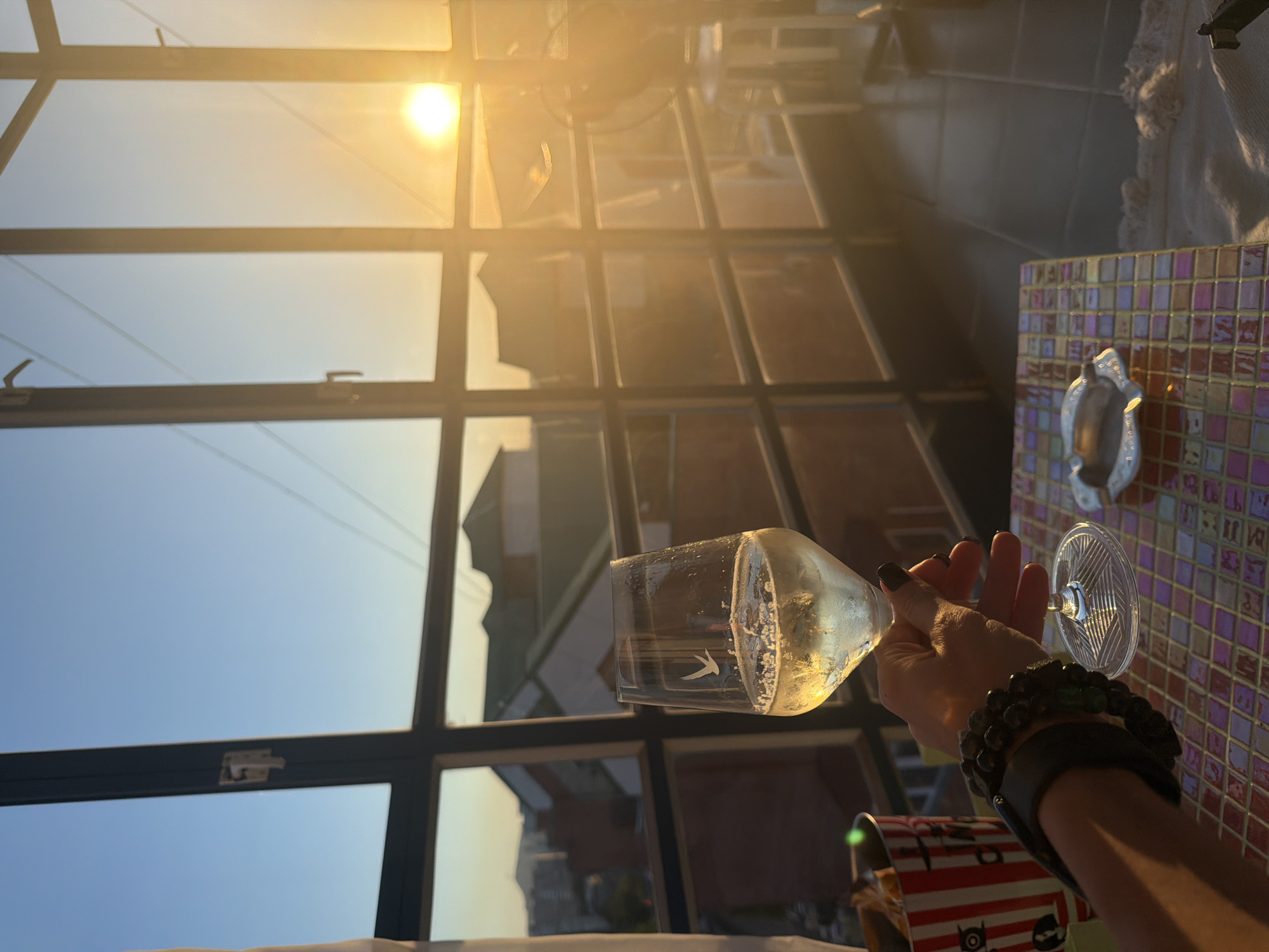 The image depicts an indoor or covered balcony space with large, dark-framed windows overlooking a residential area in Krasnoyarsk, Russia, during sunset. The sun is visible as an intense, bright orb on the right side of the frame, casting a strong golden and orange light across the scene.
In the foreground, a human right hand, featuring dark nail polish, holds a clear wine glass by its stem. The glass contains a light-colored, effervescent liquid, likely white or sparkling wine, and has a small bird logo etched onto its side. The wrist of the hand is adorned with multiple accessories, including a wide, dark-colored band and at least two beaded bracelets, one dark-colored and another with greenish-blue beads.
The hand is positioned above a small, square table with a mosaic tiled surface composed of various colored squares, including shades of pink, purple, yellow, and blue. Resting on this table is an empty, metallic, horseshoe-shaped ashtray. A red and white striped container, resembling a popcorn bucket, is partially visible in the lower-left corner of the frame.
Through the multi-paned windows, which feature visible latches suggesting they are operable, a view of suburban buildings with sloped roofs and distant hills or mountains is visible under a sky that transitions from light blue at the top-left to a warm orange and yellow near the horizon. The floor of the immediate space is covered with dark tiles. A white stool or piece of furniture is partially visible on the far right side of the image, close to the brightly shining sun.