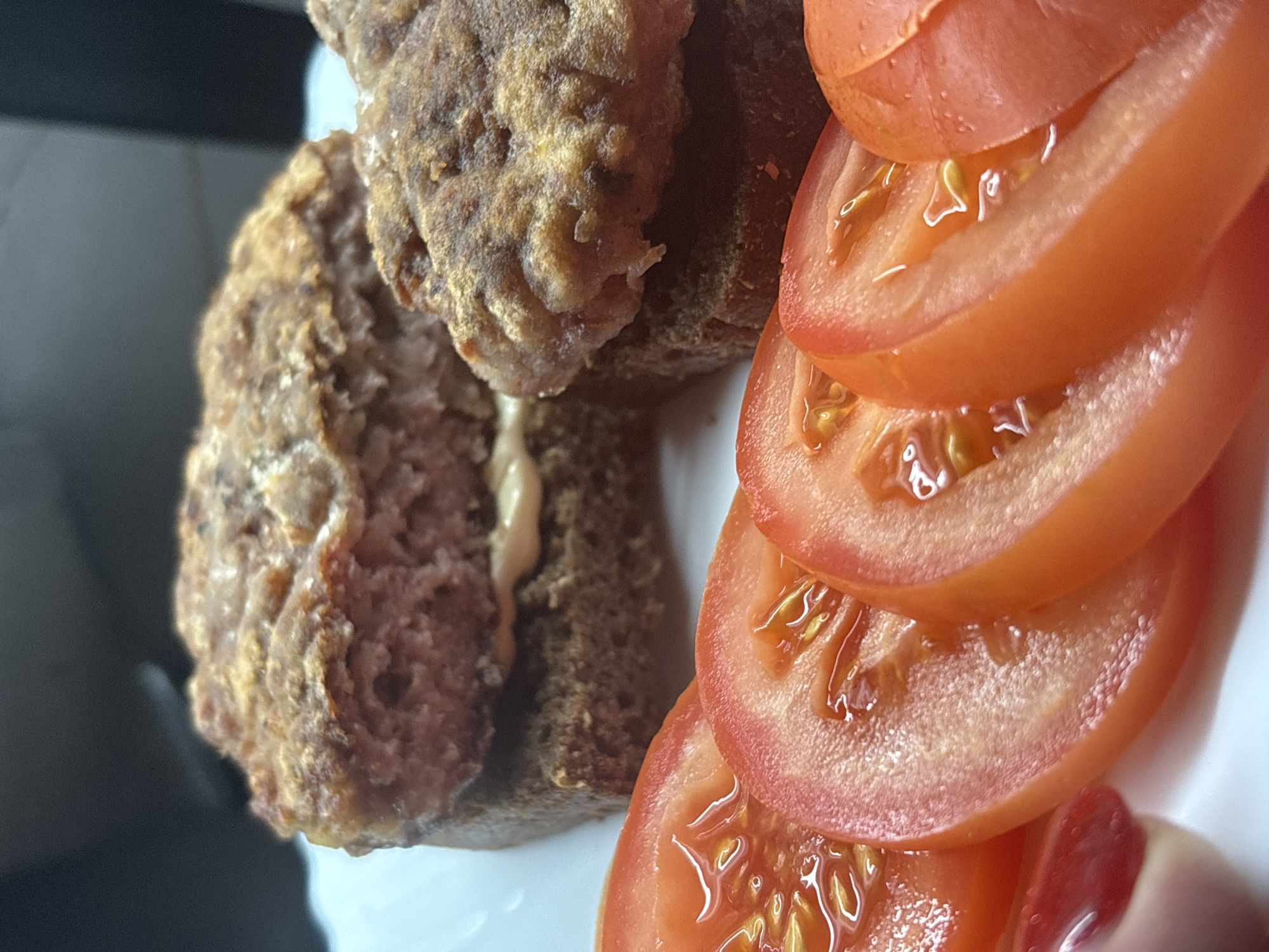In Sochi, Russia, on a white plate, a burger is served with sliced tomatoes. The burger is made with two patties, each on a slice of dark bread. The patties are thick and appear to be fried. The bottom patty has a white sauce on it. Sliced tomatoes are arranged in front of the burger, with their glossy, seedy interiors visible. A person's hand with red nail polish is partially visible on the left side of the image. The background is dark and out of focus.