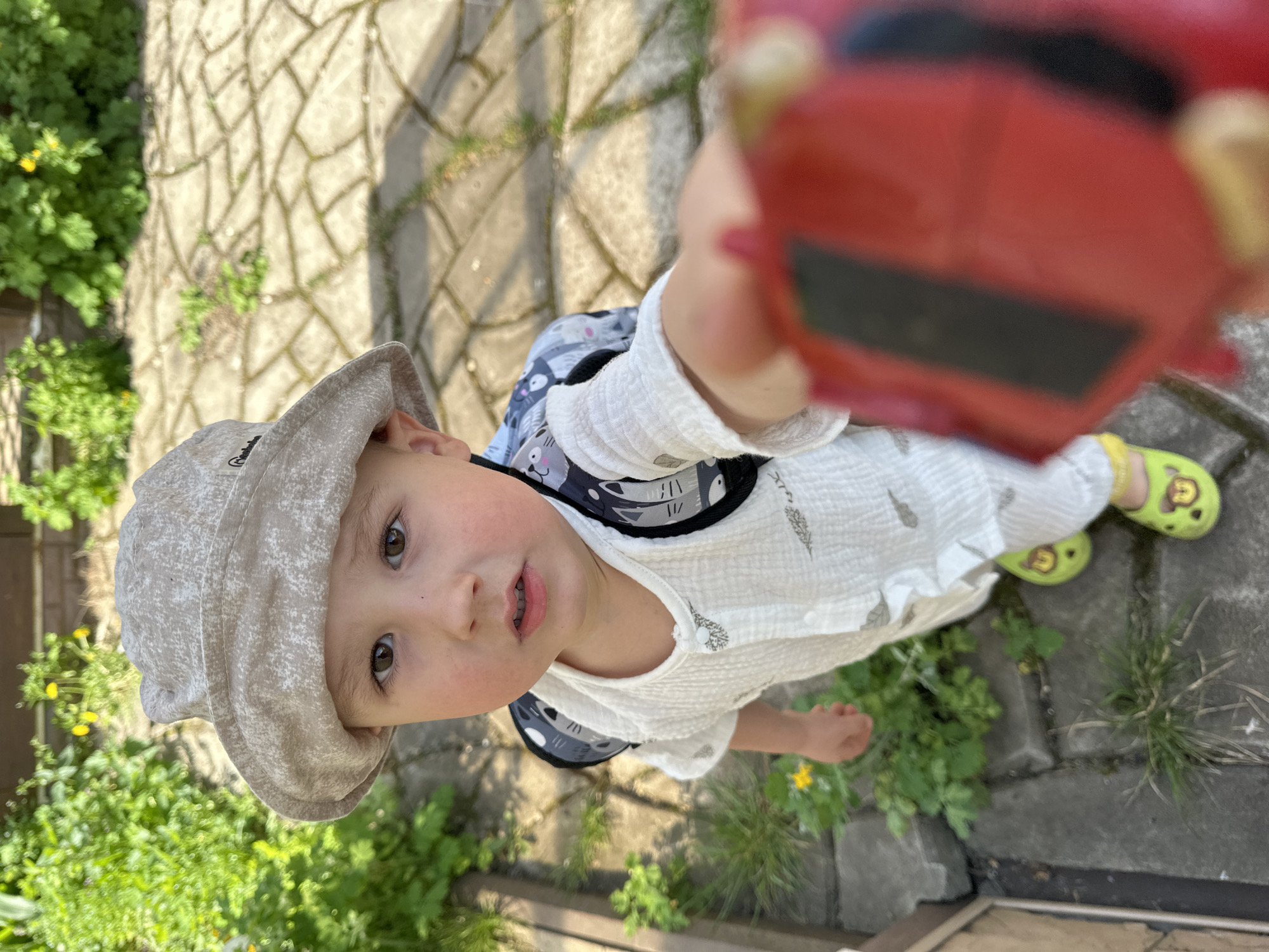 A child stands on a stone-paved surface, holding a blurry, red object up to the camera. The child is wearing a tan bucket hat, a white shirt with a pattern of feathers, and a backpack with a gray and white cat print. The child is also wearing green Crocs-style shoes. The environment includes patches of green vegetation. There is no concrete information about the location in the city of Nizhny Novgorod, Russia.